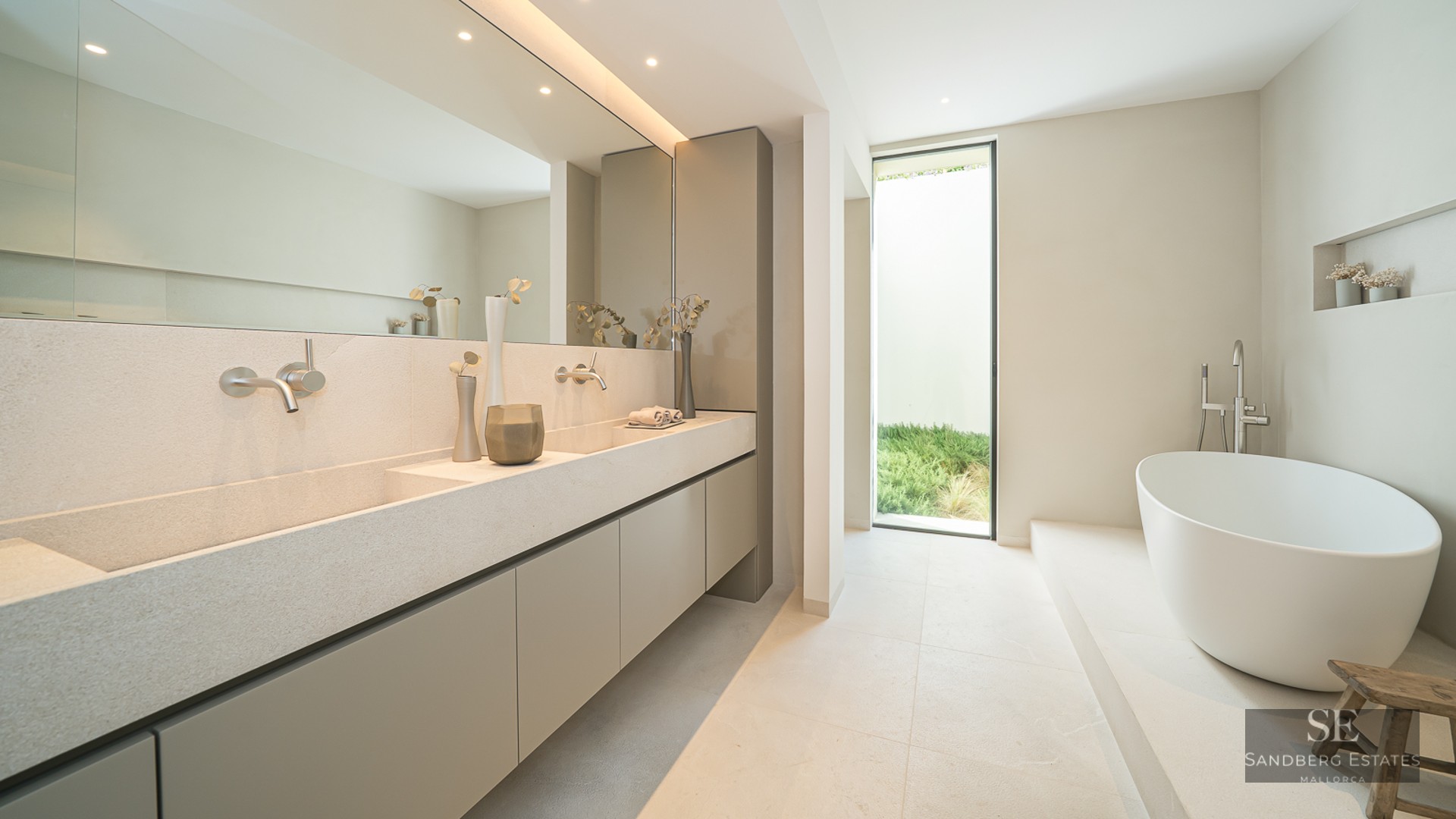 Minimalist bathroom with a stone double vanity, large mirror, freestanding white tub, and a window to a garden.
