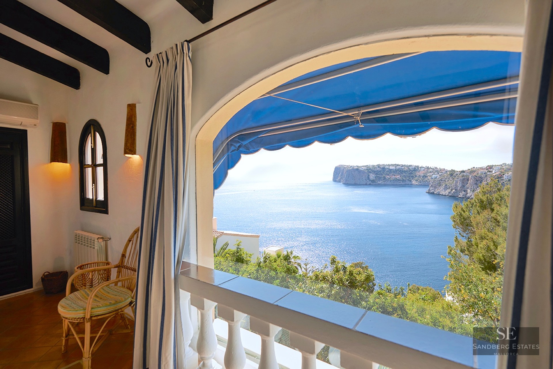 Covered terrace with wicker chair, blue awning, and panoramic view of the blue Mediterranean sea and rocky cliffs.