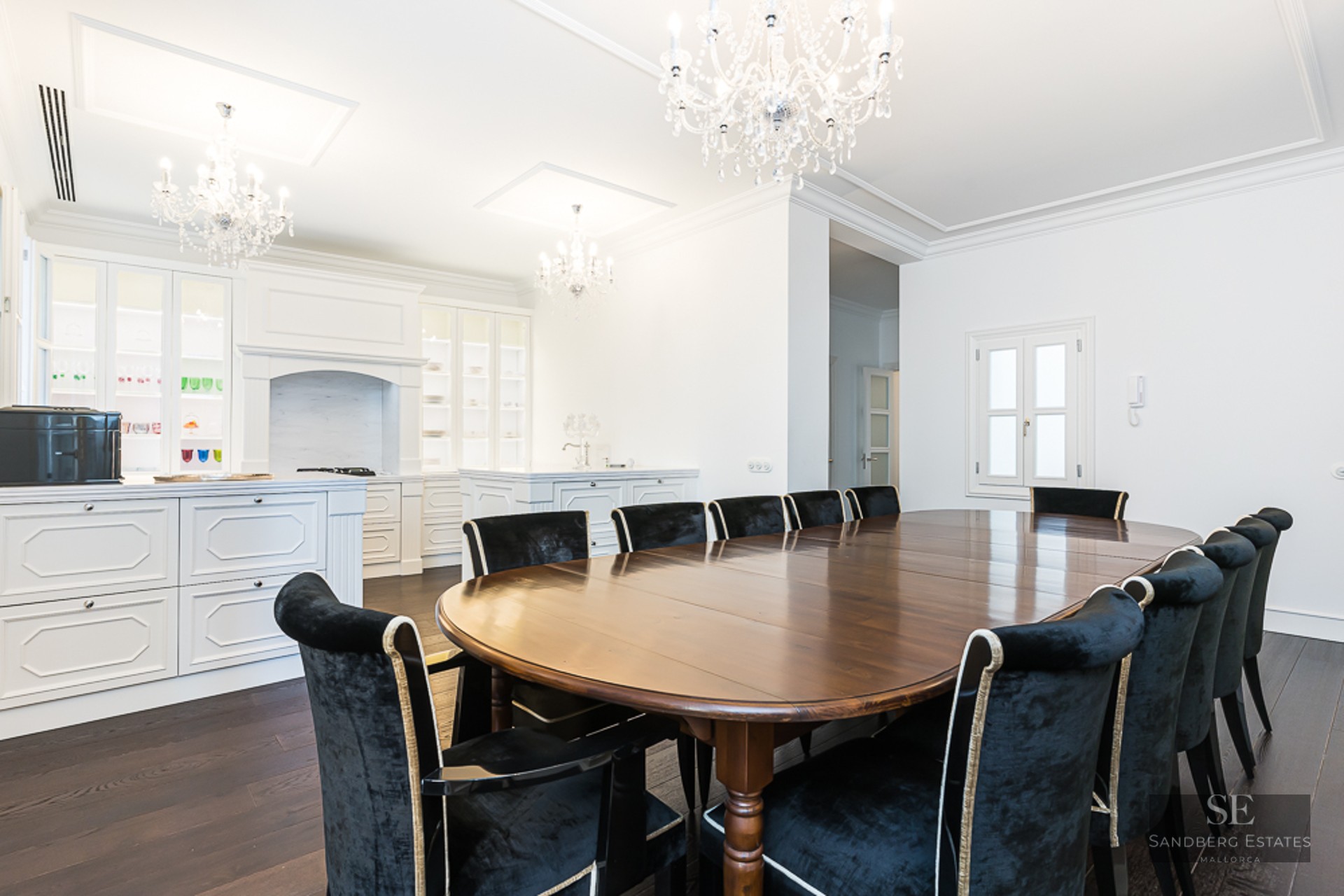 Large oval dark wood dining table surrounded by black velvet chairs under ornate crystal chandeliers in a white room.