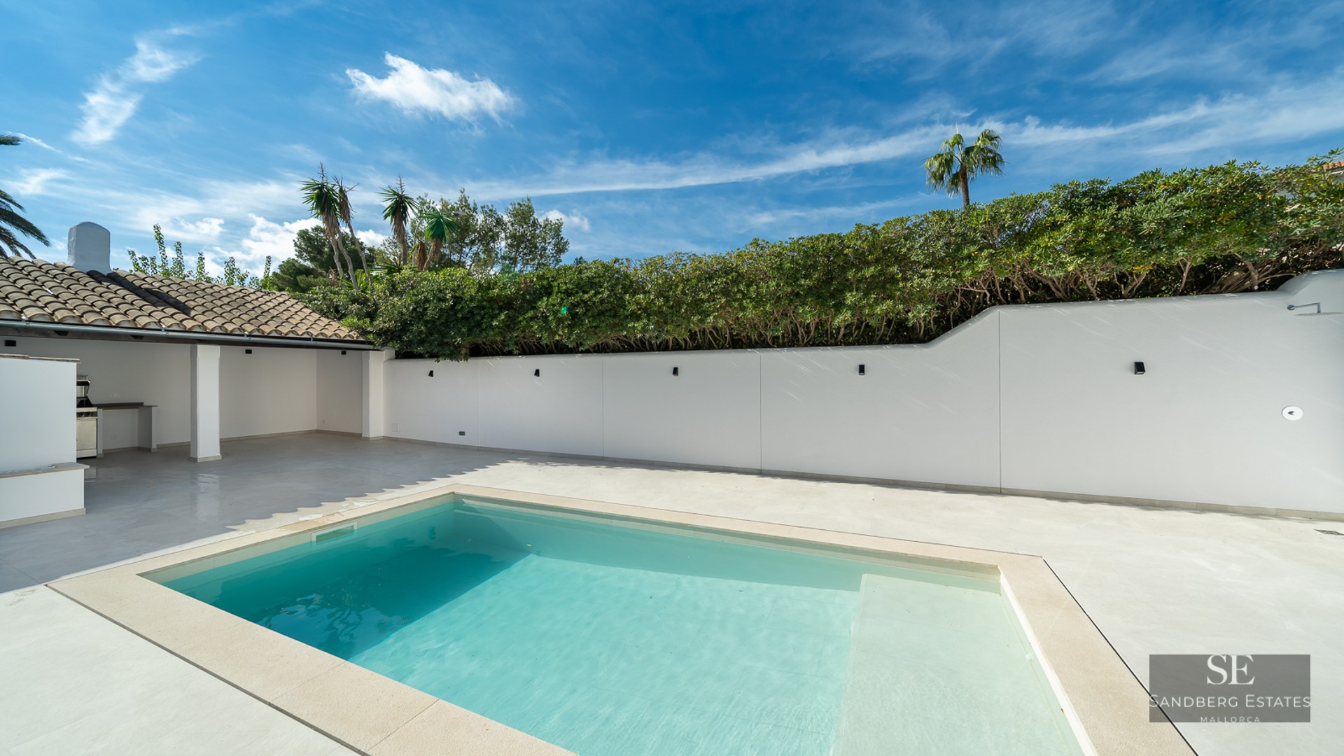Turquoise swimming pool on a white stone terrace with a privacy wall, lush hedges, and an outdoor kitchen area.