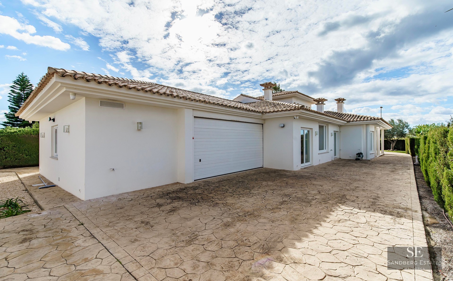 Spacious stamped concrete driveway with a white garage door in a Mediterranean style villa under a bright cloudy sky.