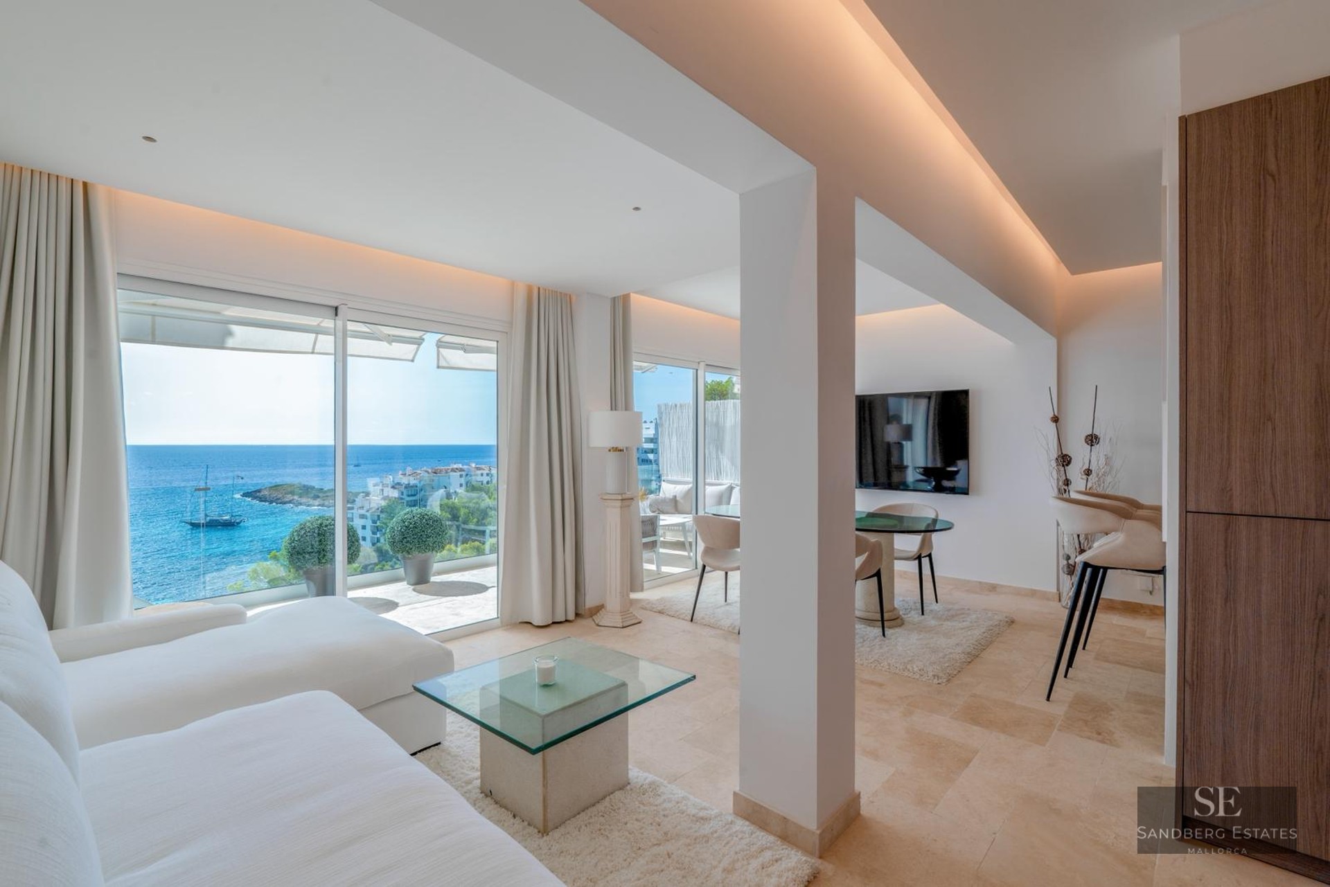 Bright modern living room with large glass doors opening to a Mediterranean sea view, featuring white sofas and stone floors.