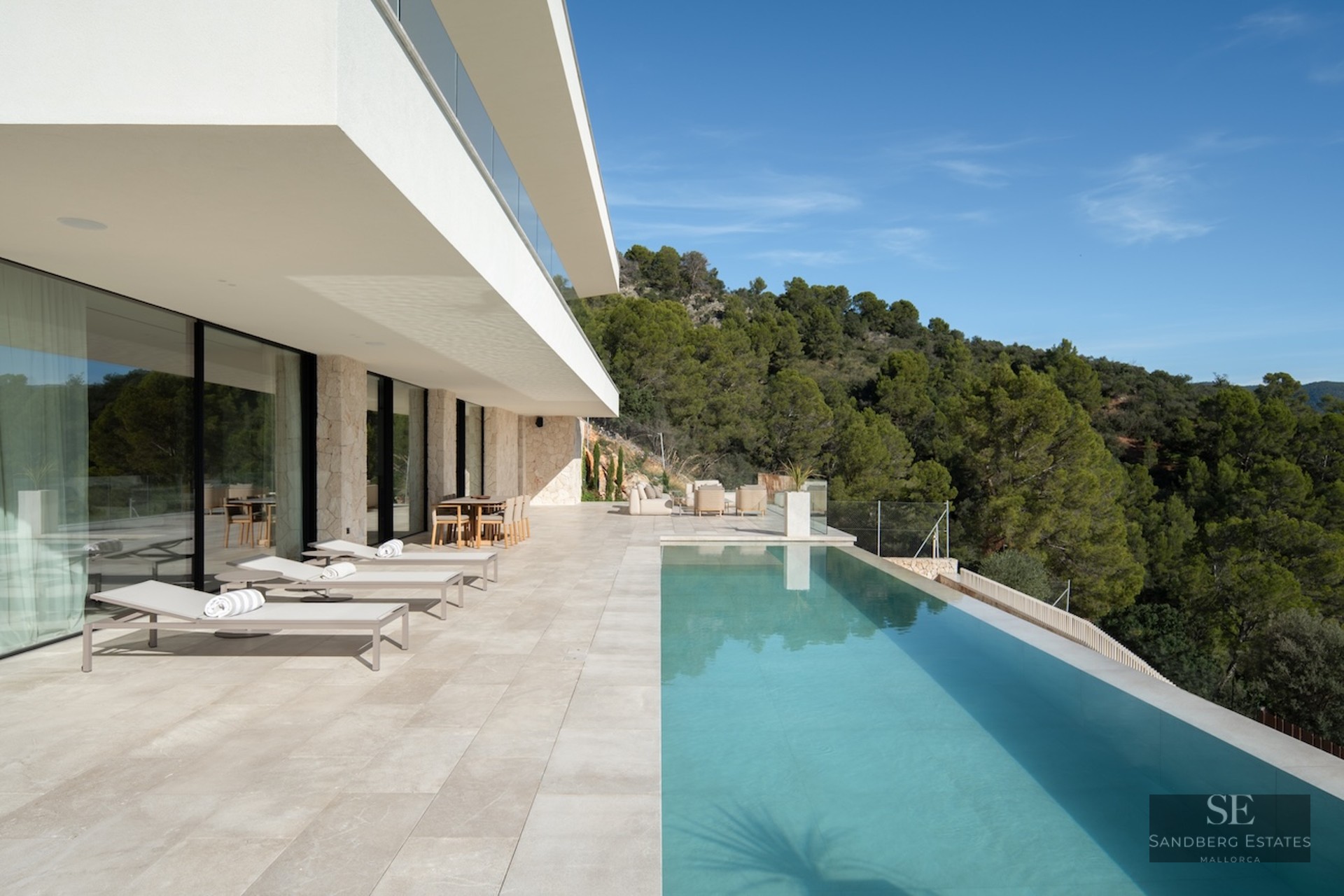 A spacious white stone terrace featuring an infinity pool, sun loungers, and views of a pine forest under a blue sky.