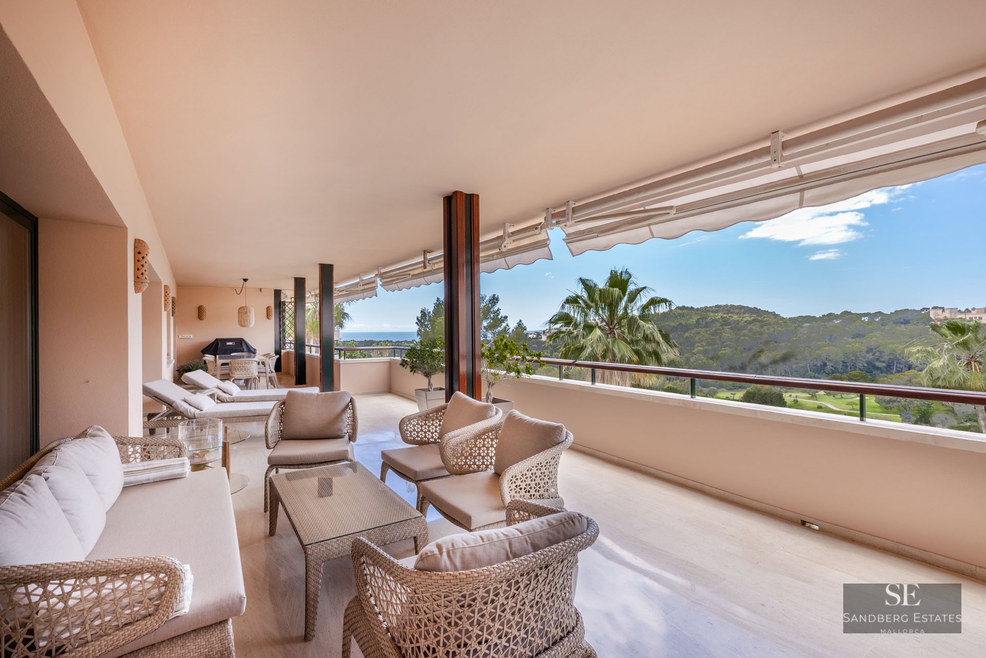 Grand balcon couvert avec mobilier en osier offrant une vue sur une vallée verdoyante et la mer par temps clair.