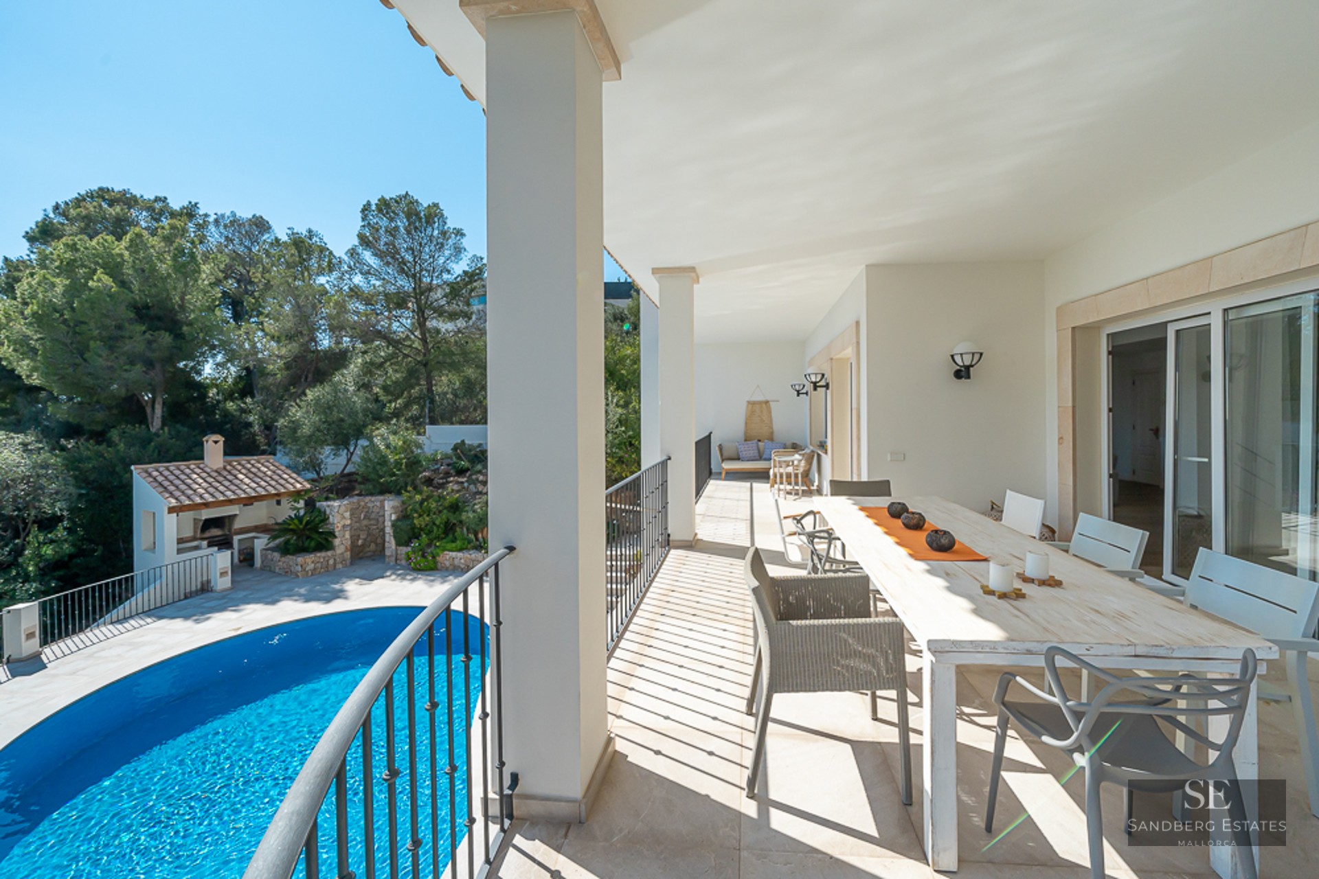 Terrasse couverte blanche avec grande table à manger et vue sur une piscine bleue et de la verdure.