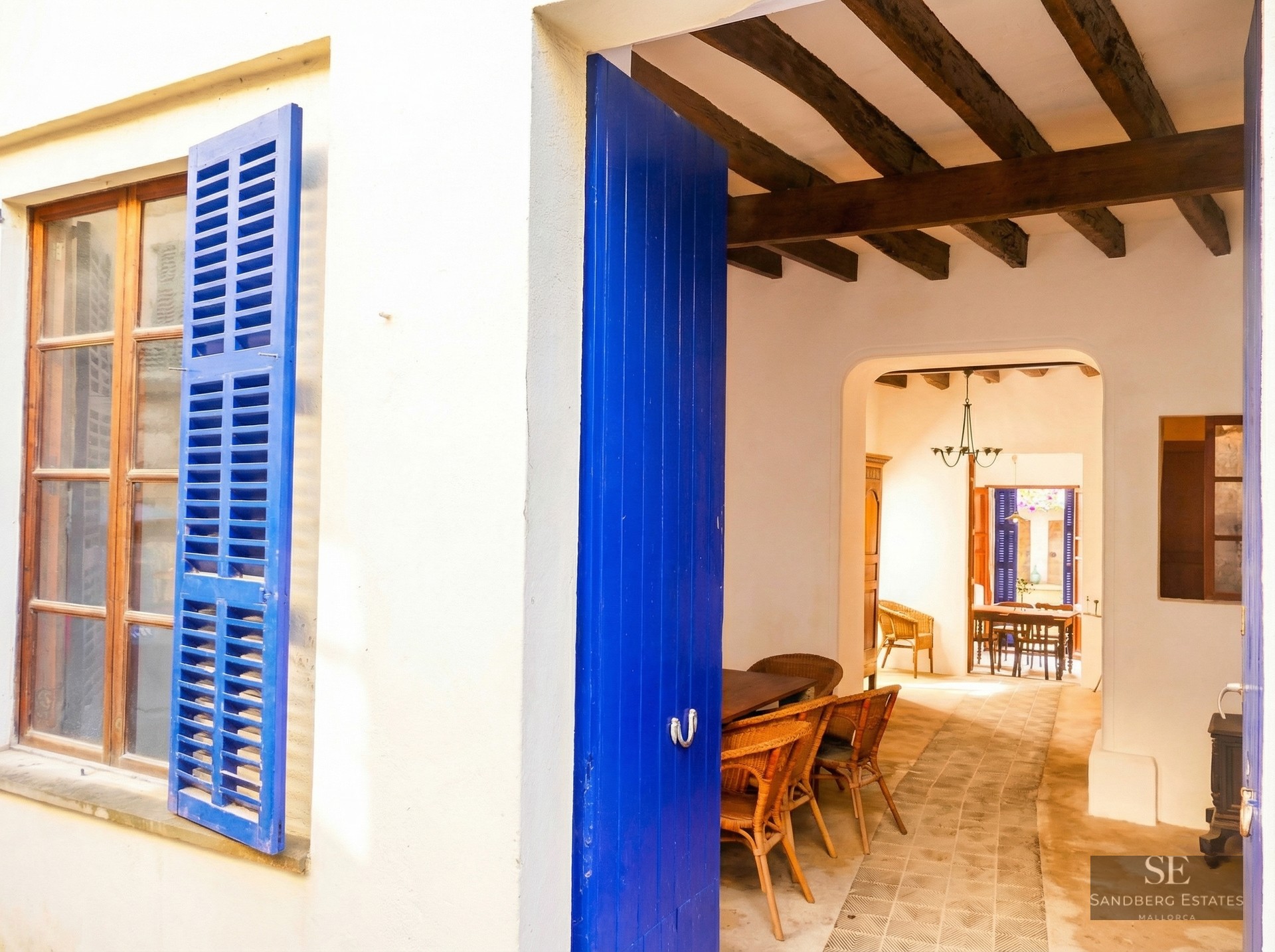 White exterior wall with bright blue shutters and open door leading to a rustic interior with wooden beams.