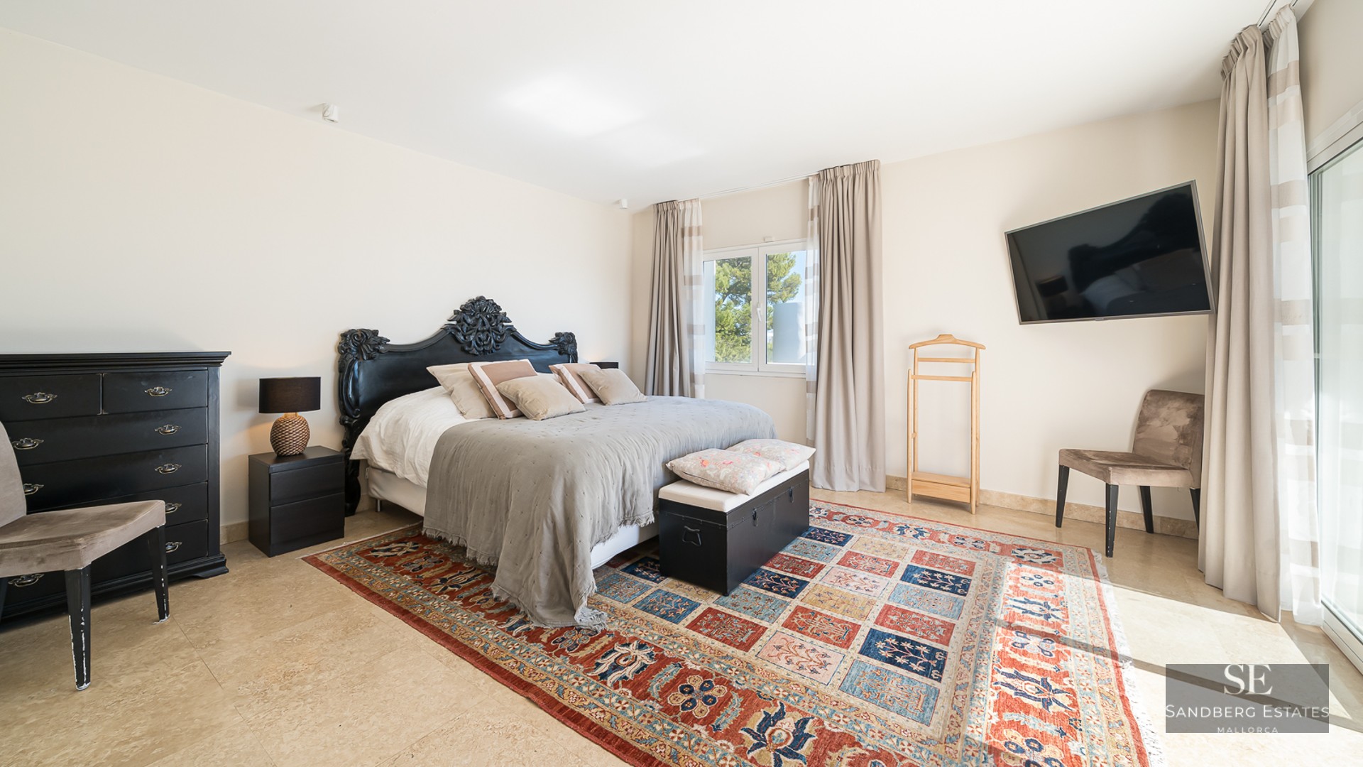 A bright master bedroom with an ornate black headboard, a colorful patterned rug, and travertine tile floors.