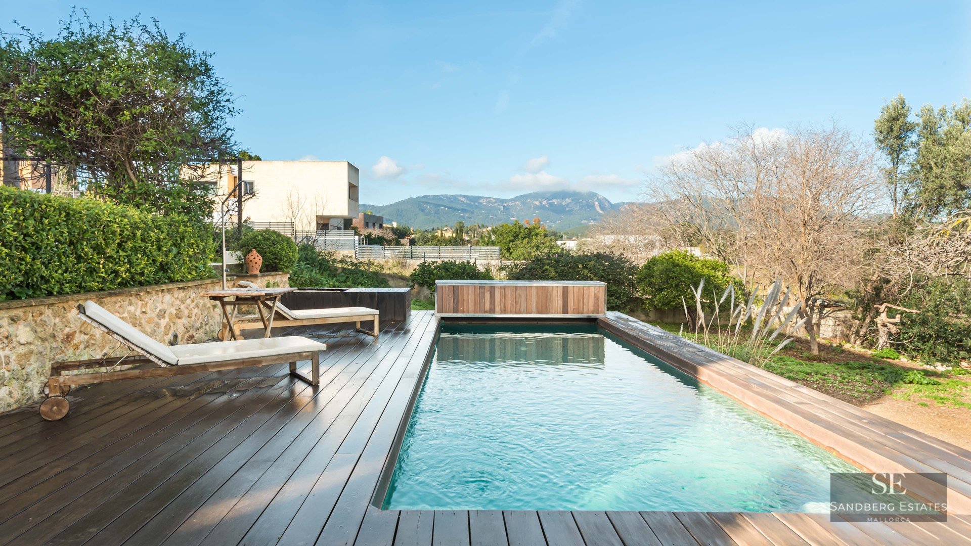 A rectangular swimming pool with a dark wood deck, white sun loungers, and a scenic mountain backdrop.