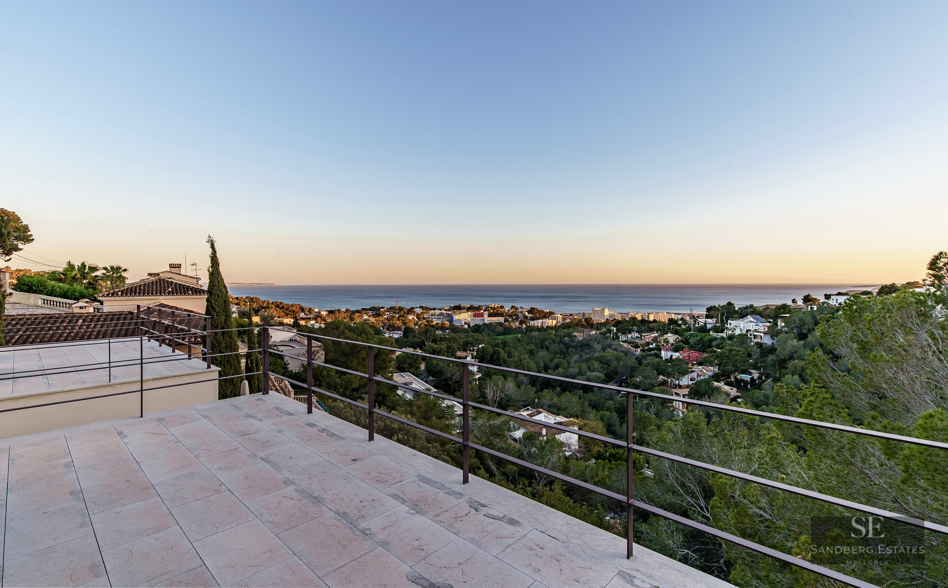 Wide stone terrace with dark metal railings overlooking a coastal town and the blue sea at sunset.