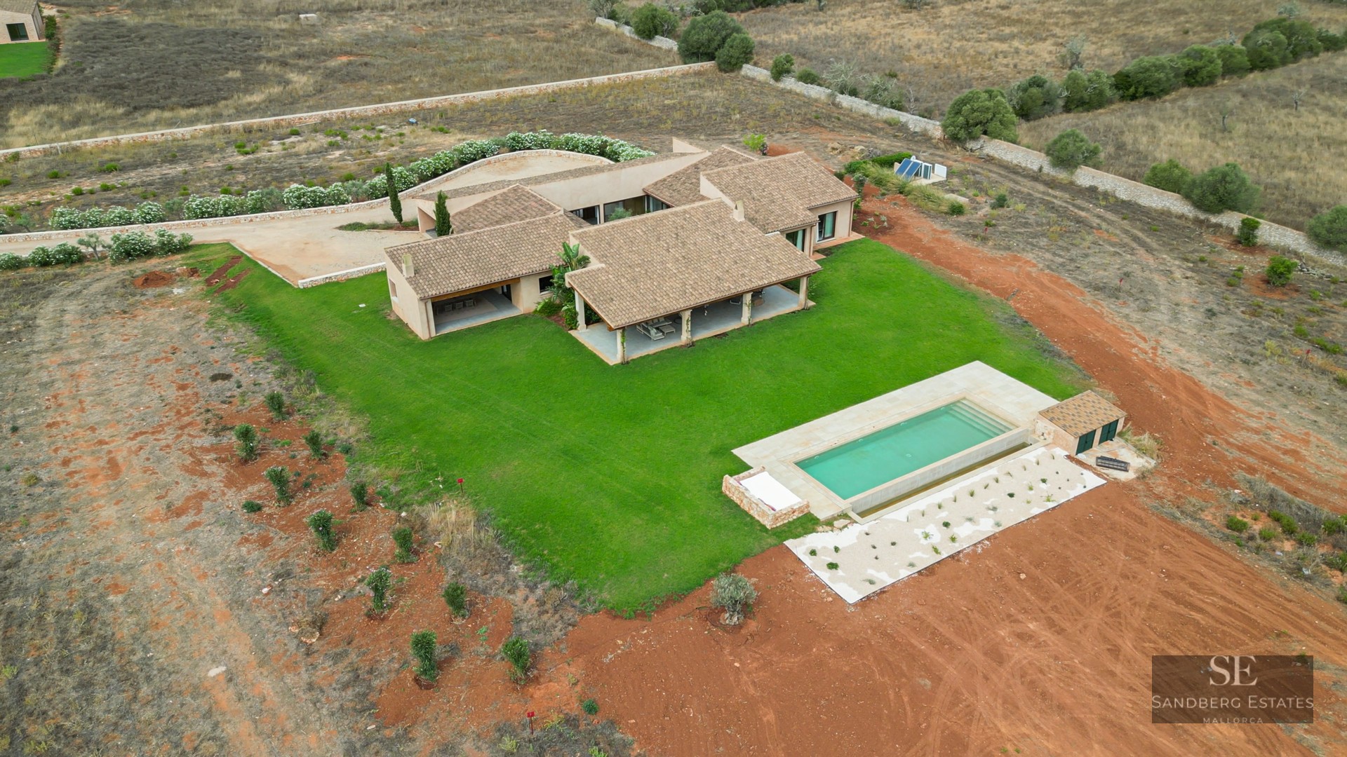 Aerial drone shot of a modern villa with terracotta roof, green lawn, and rectangular pool in the countryside.