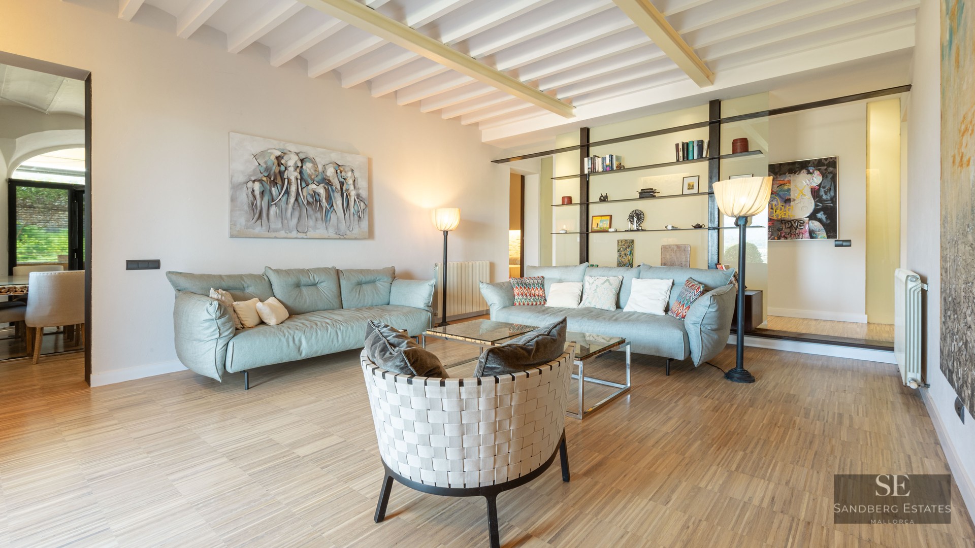 Spacious modern living room with light blue sofas, a woven designer armchair, and white exposed ceiling beams.