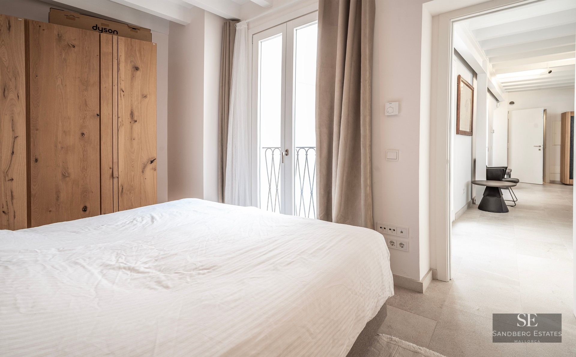 Chambre lumineuse avec lit blanc, grande armoire en bois et portes-fenêtres donnant sur un balcon.