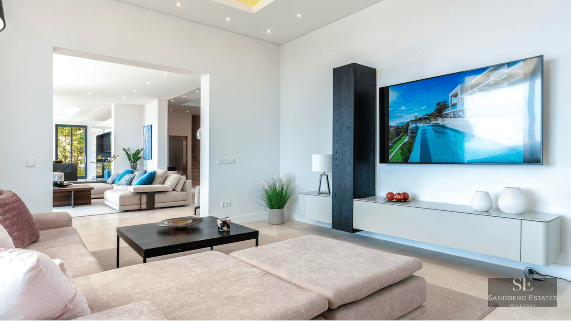 Spacious modern living room featuring a large wall-mounted TV, beige sofas, and minimalist dark wood furniture.