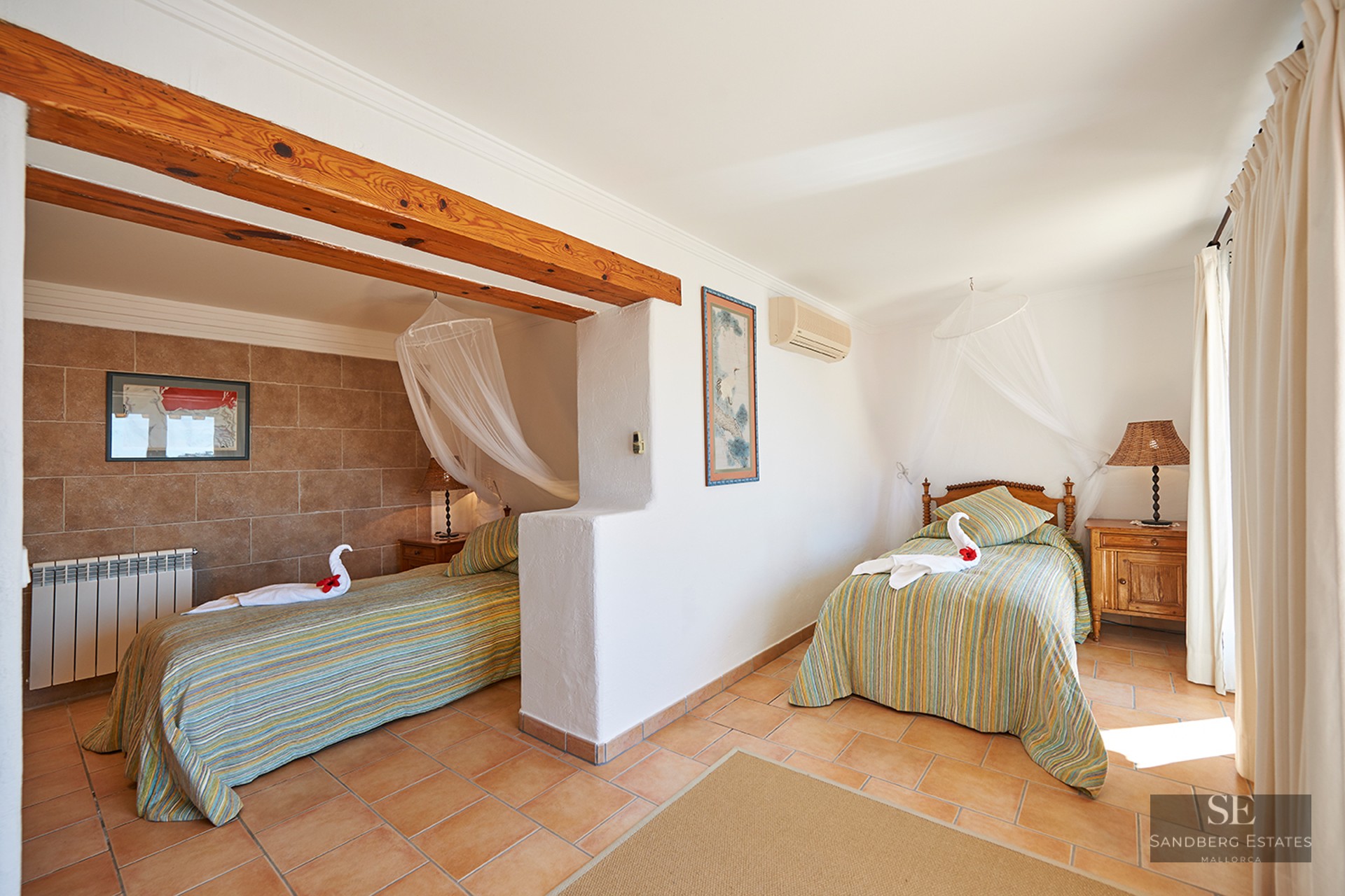 Bright bedroom featuring two twin beds with mosquito net canopies, exposed wooden beams, and terracotta tile floors.