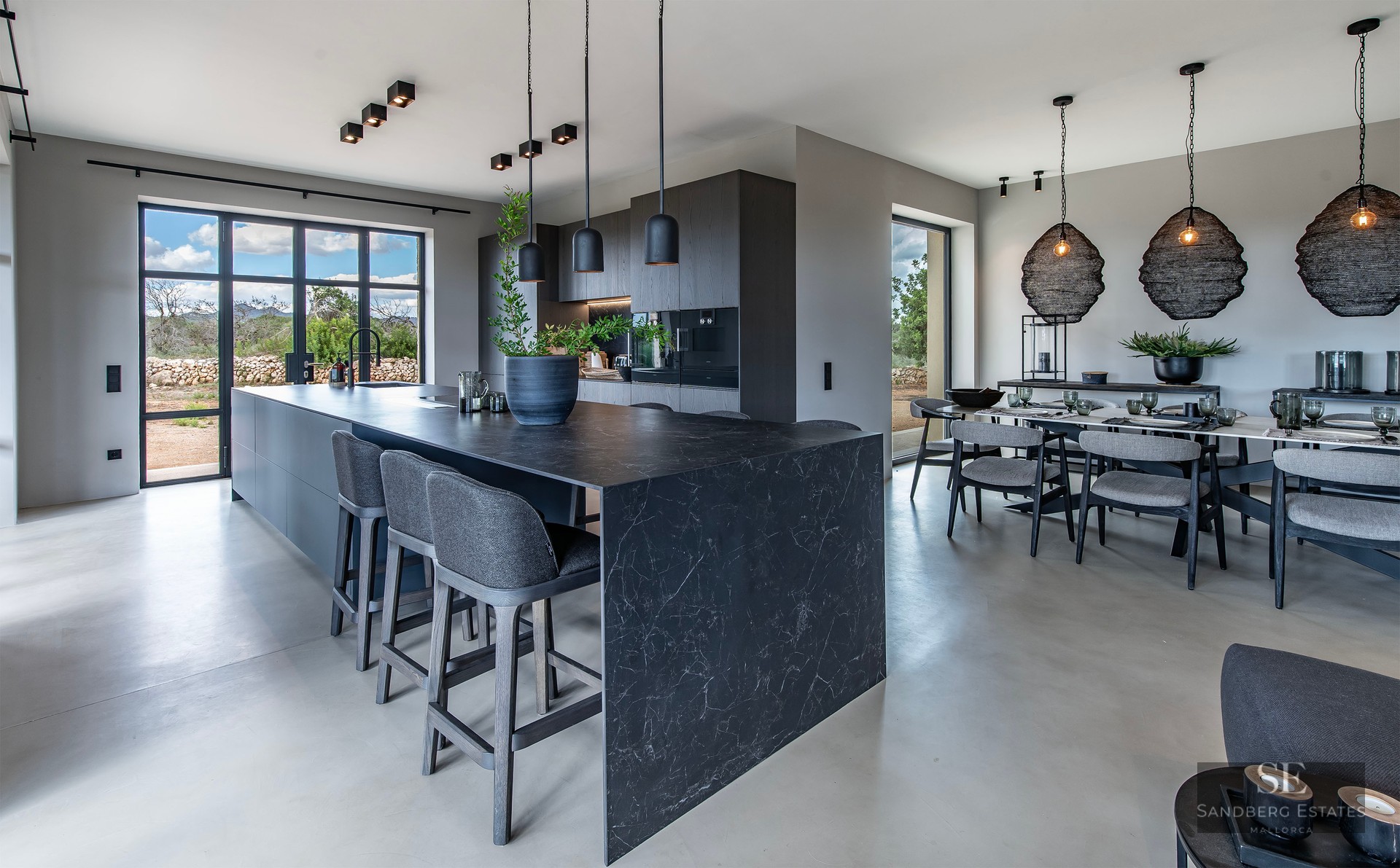 Modern open-plan kitchen with a dark marble island, grey stools, and a dining table under black designer pendant lights.
