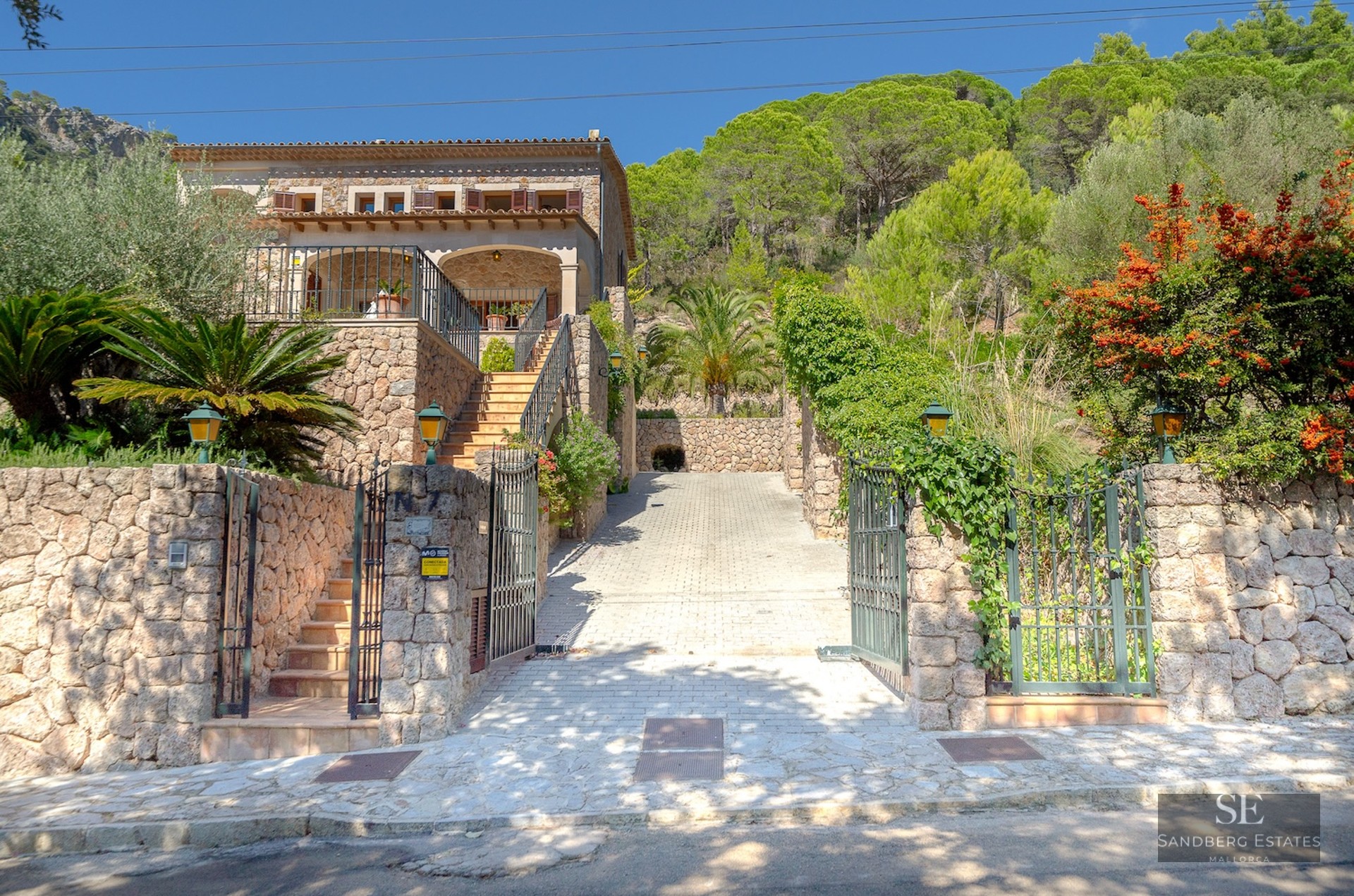 Two-story traditional stone house with iron gates, a paved driveway, and lush green trees against a bright blue sky.