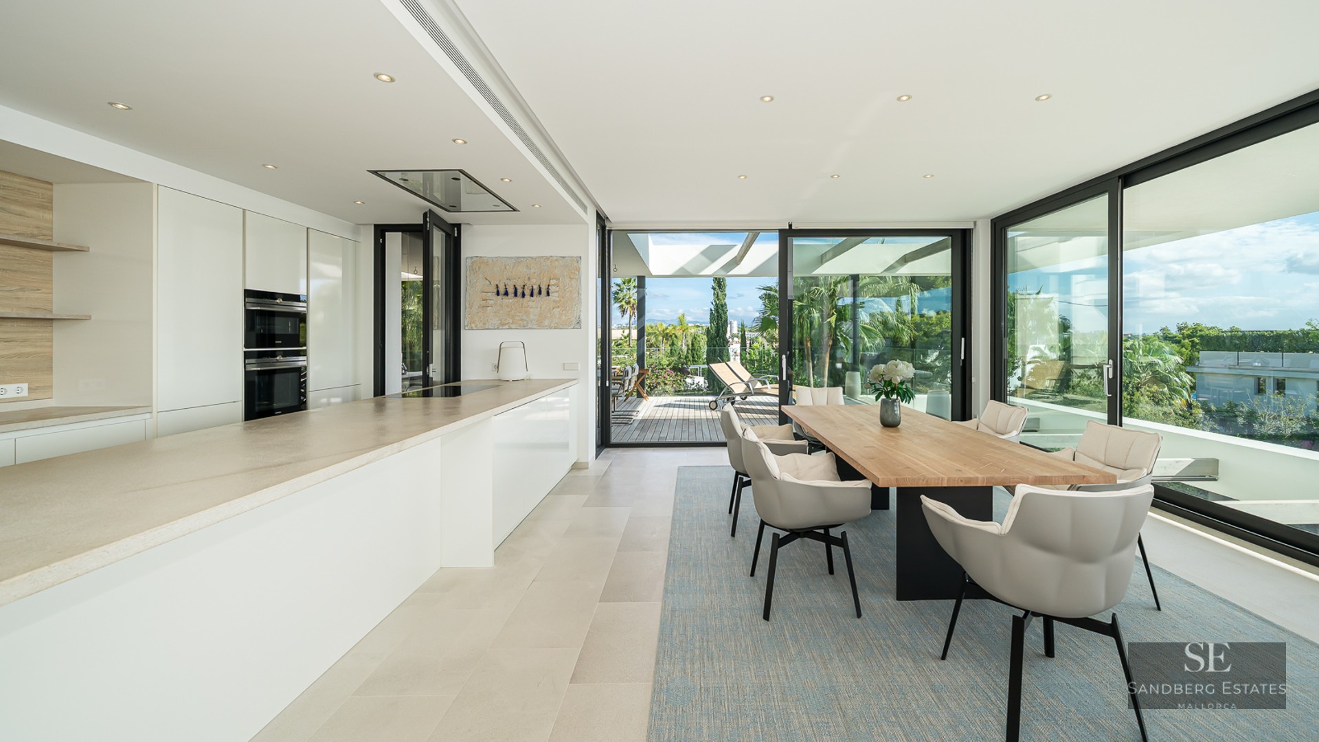 Modern dining room with a large wooden table, grey chairs, and a sleek white kitchen with floor-to-ceiling glass doors.