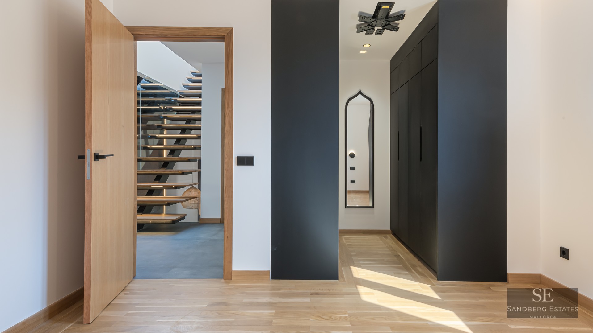 Modern hallway with light wood floors, black built-in closets, and an open door viewing a floating wooden staircase.