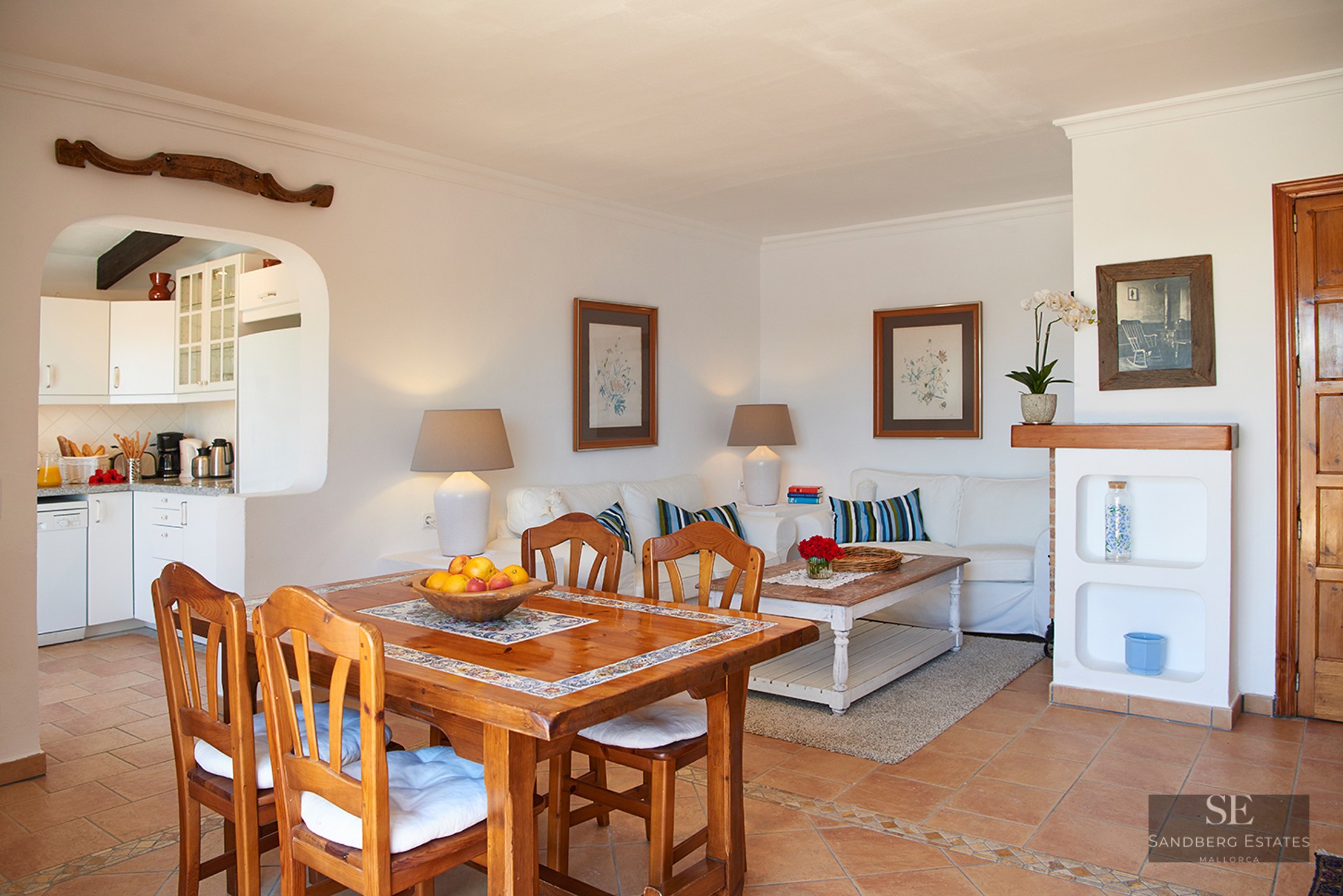 A bright living and dining space with terracotta tile floors, a wooden dining table, and a white sofa near a kitchen arch.