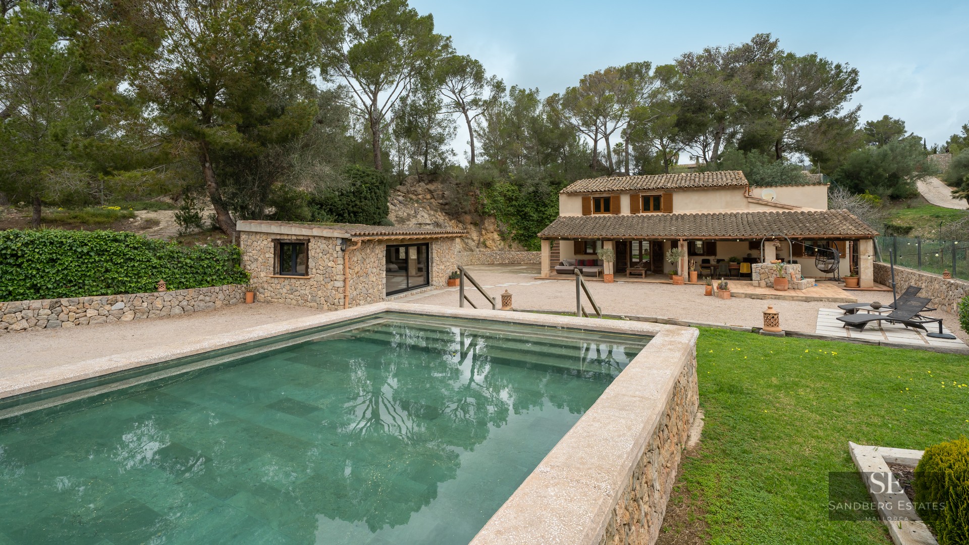 Outdoor swimming pool with stone borders in front of a rustic Mediterranean villa surrounded by pine trees.