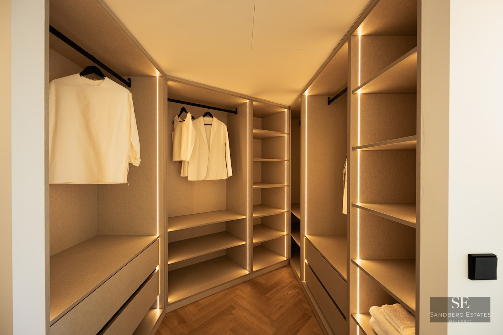 Modern walk-in closet with warm LED strip lighting, light wood cabinetry, and herringbone parquet flooring.