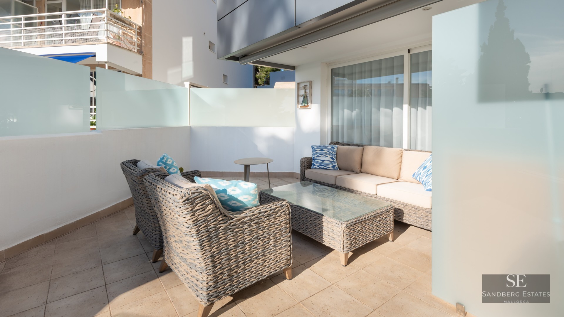 Terrasse moderne avec canapés en osier gris, coussins beige et bleus sur un patio ensoleillé avec parois en verre dépoli.