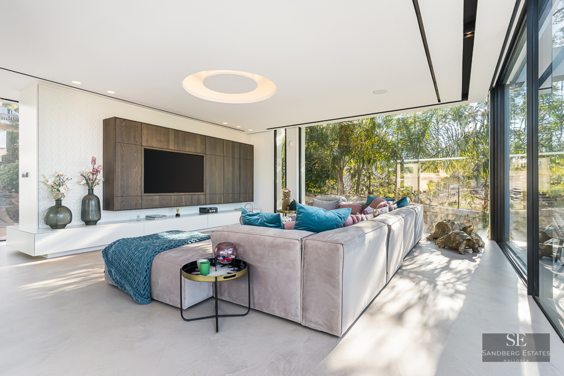 A bright contemporary living room with a large sectional sofa, wall-mounted TV, and floor-to-ceiling garden windows.