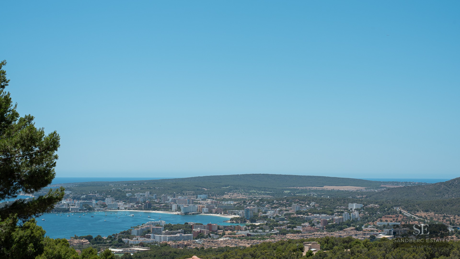 Panoramic elevated view of a blue Mediterranean bay, coastal town, and green hills under a clear blue sky.