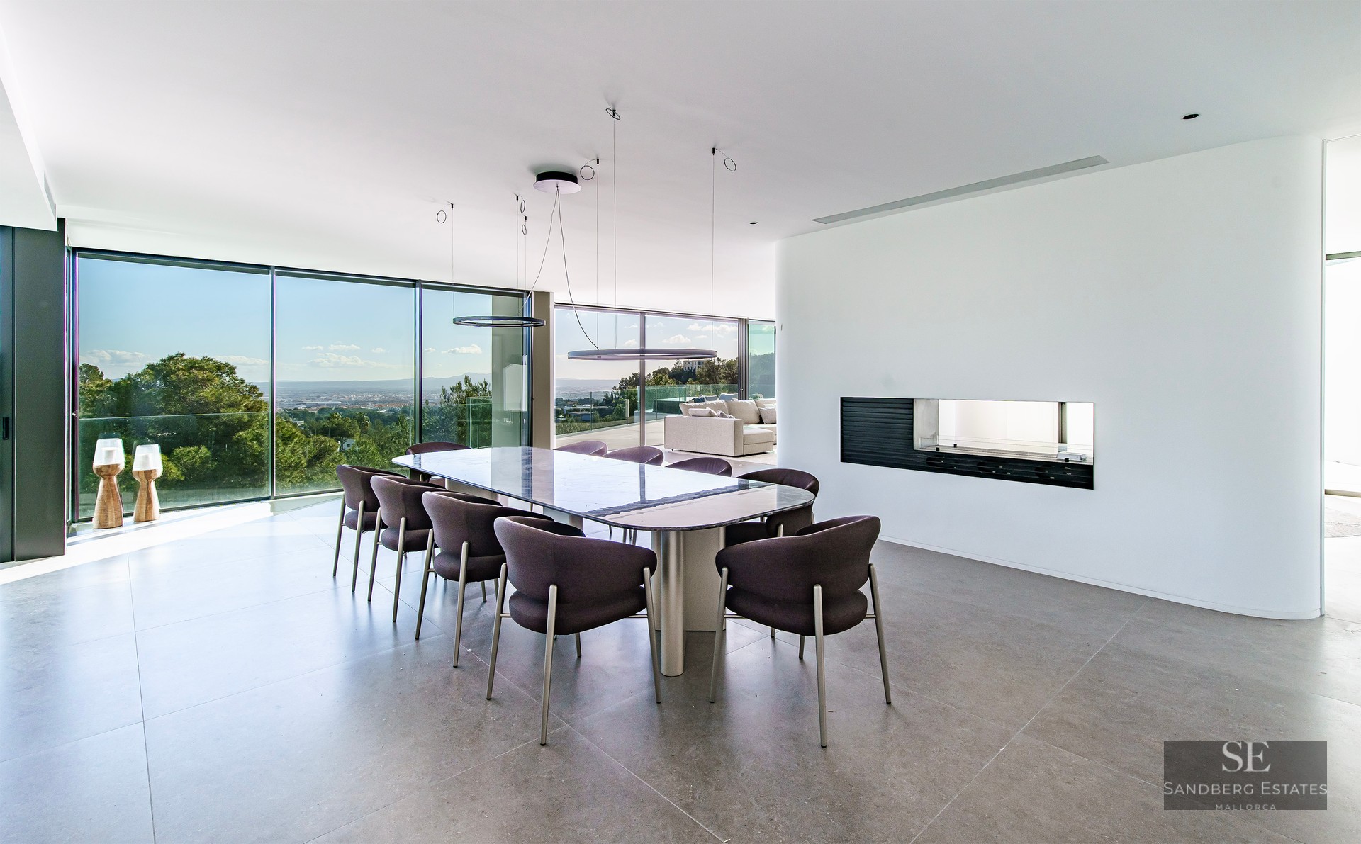 A modern dining room with a long table, dark grey chairs, and floor-to-ceiling windows overlooking a lush landscape.