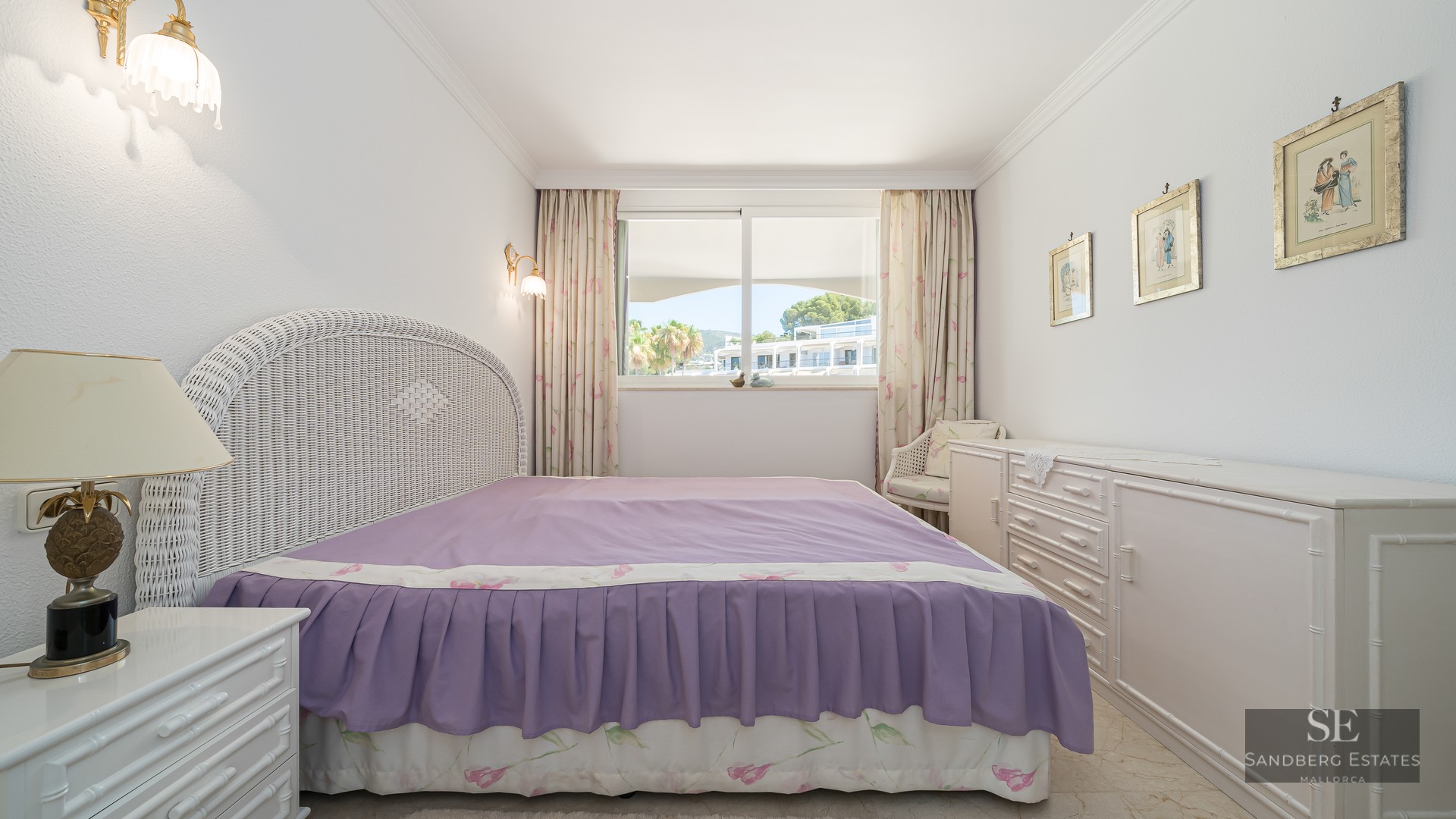 Bedroom featuring a white wicker headboard, purple bedspread, and a large window overlooking a sunny courtyard.