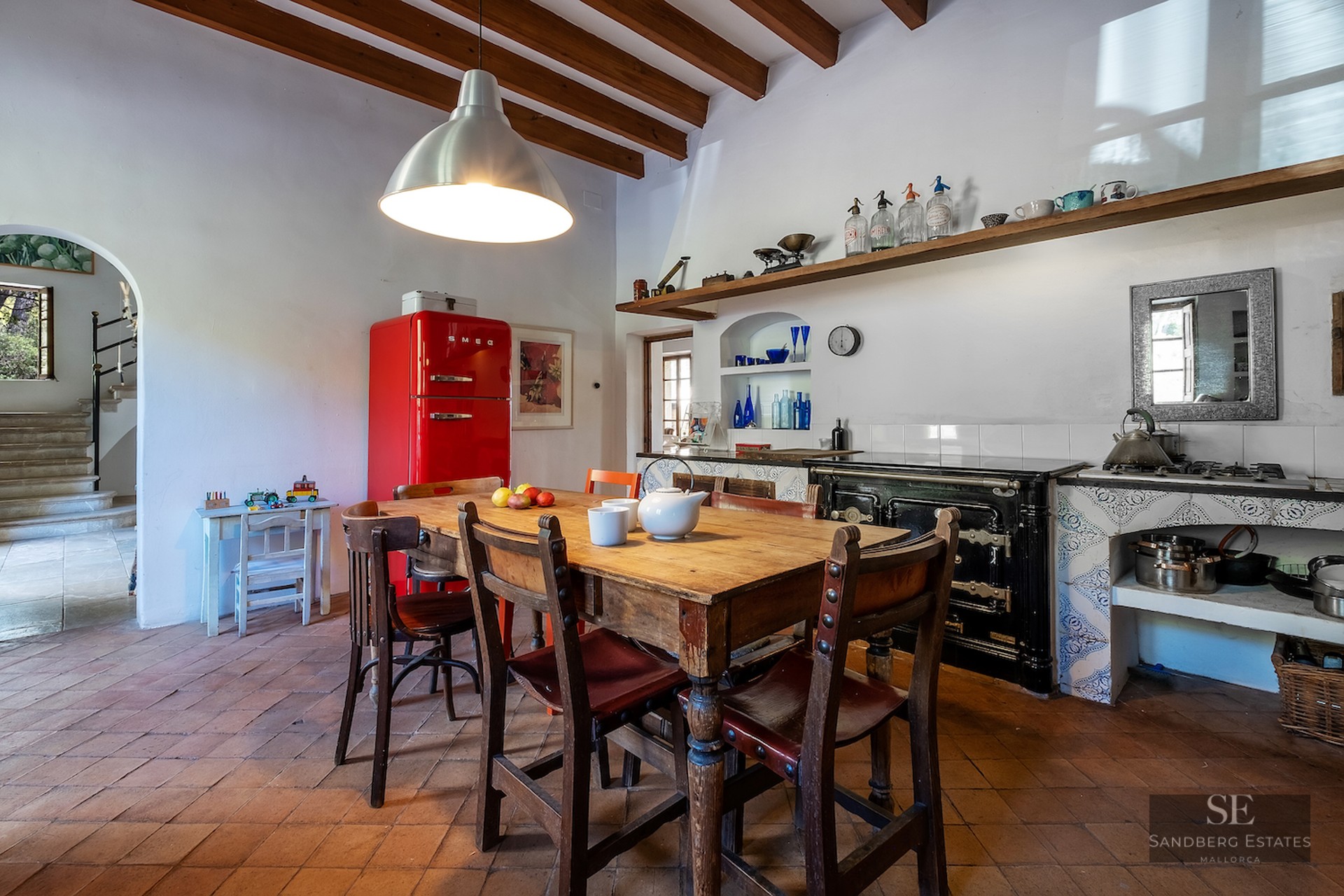 Traditional kitchen with terracotta floors, wooden beams, a rustic dining table, and a vibrant red retro refrigerator.