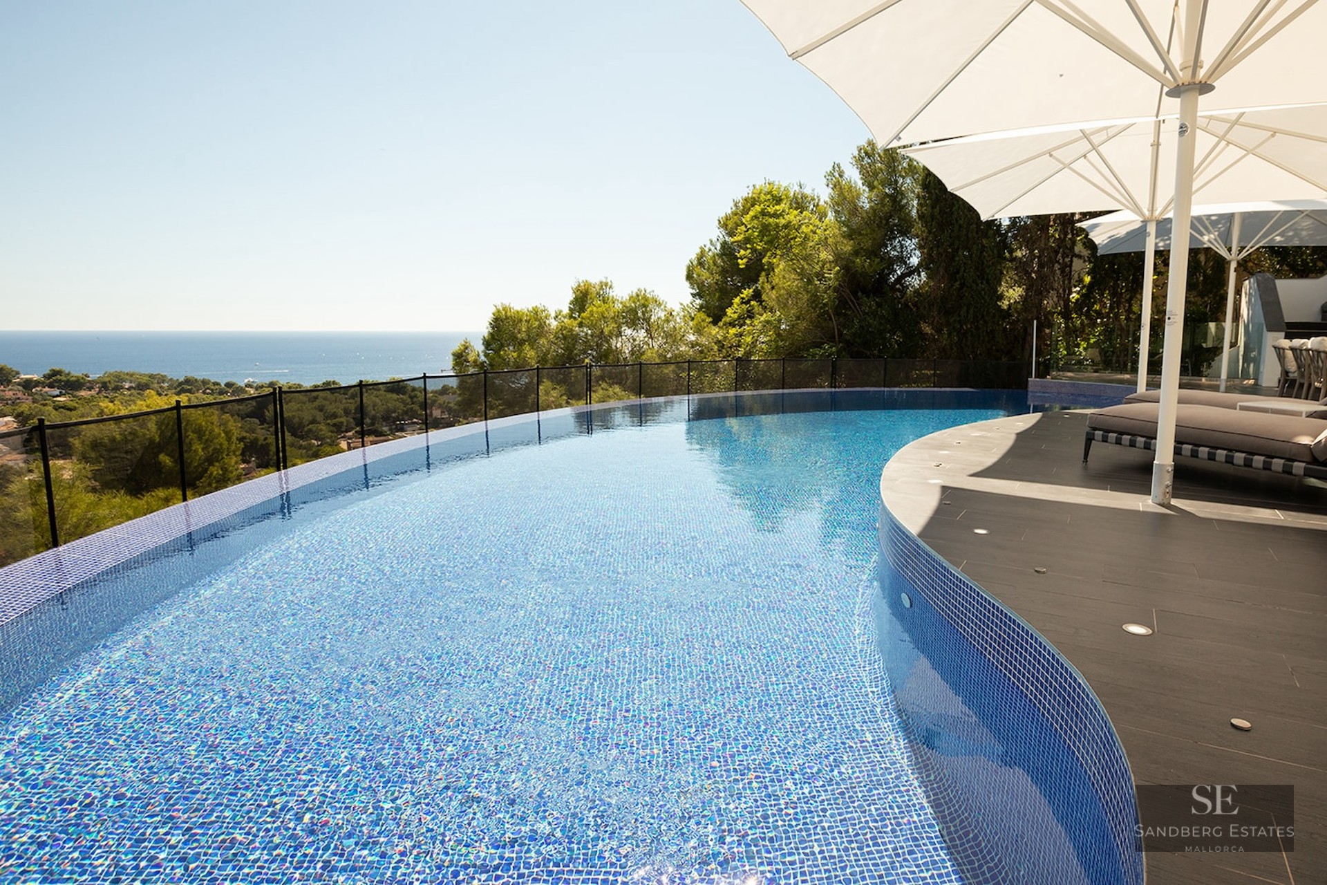 Curved infinity pool with blue mosaic tiles overlooking a lush green landscape and the blue ocean under a clear sky.