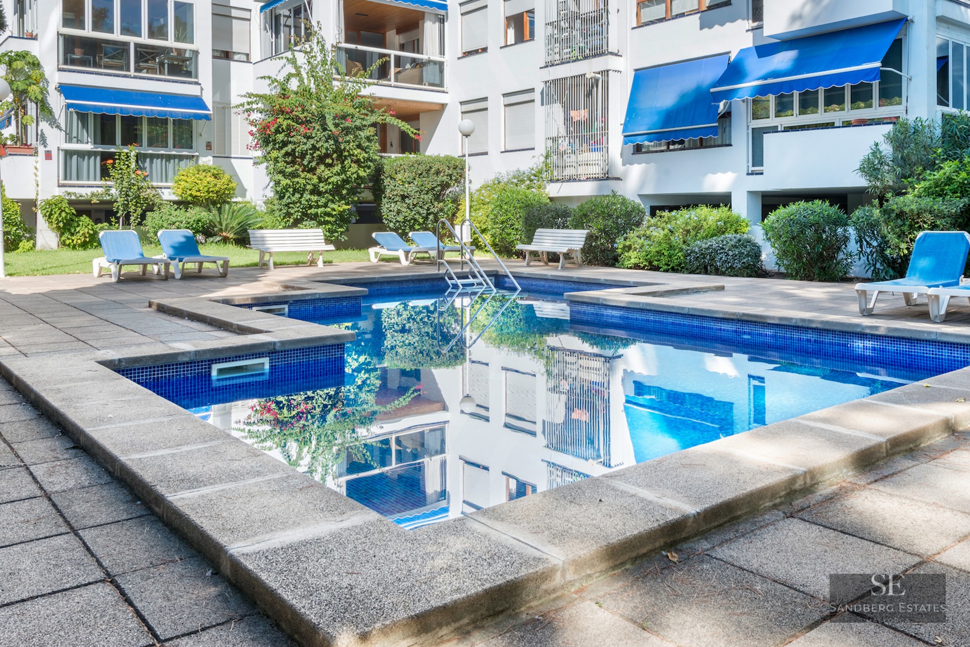 Rectangular blue pool surrounded by a stone terrace with loungers in front of a white apartment building.