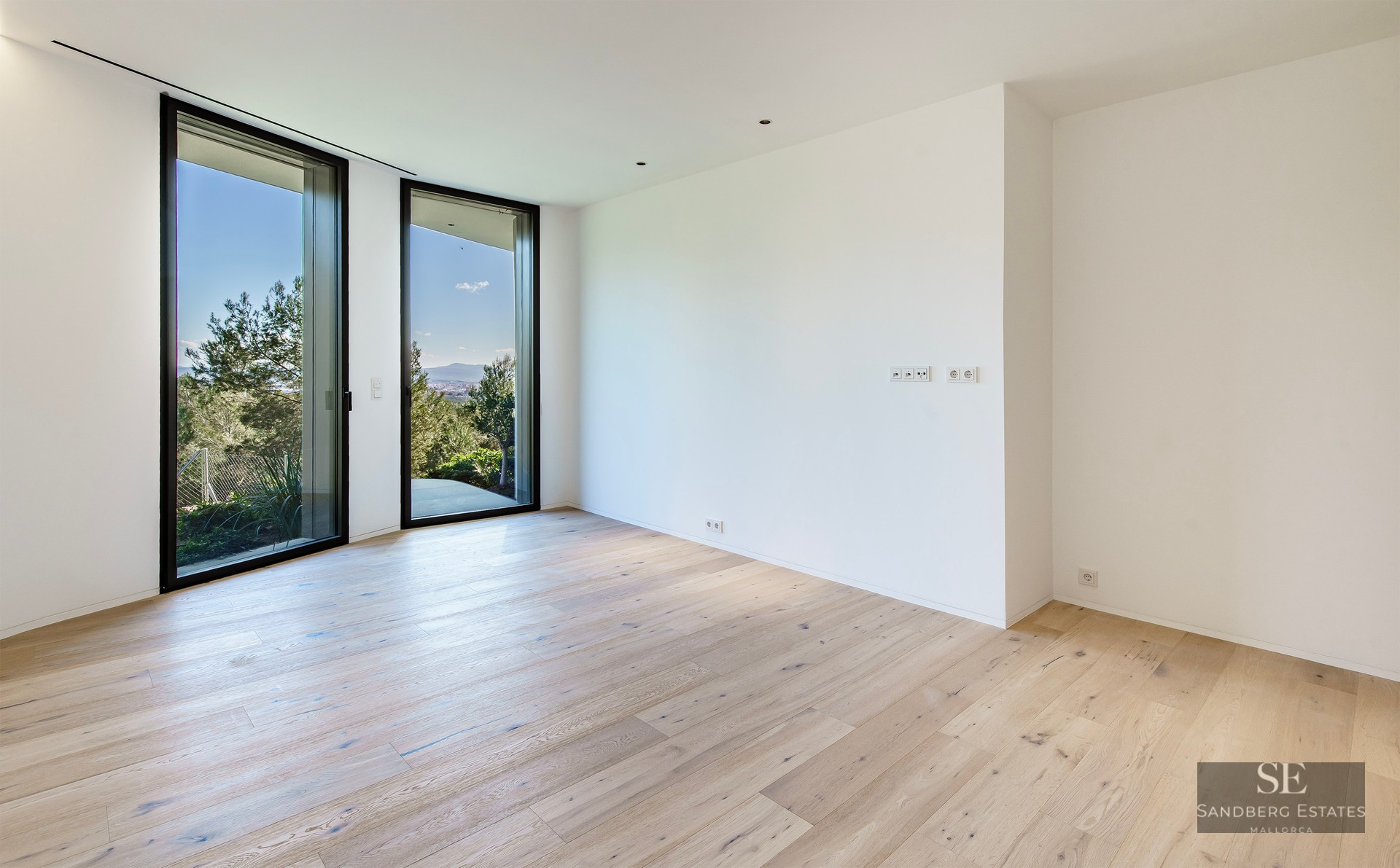 An empty room with light wood floors, white walls, and two large floor-to-ceiling windows looking out onto green trees.