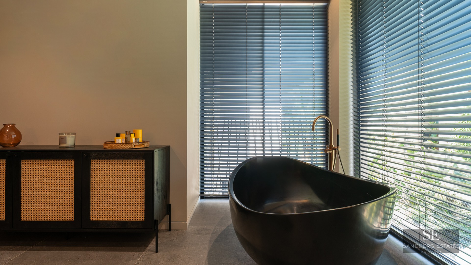 Luxury bathroom featuring a black oval bathtub, gold faucet, and a black rattan sideboard next to windows with blinds.