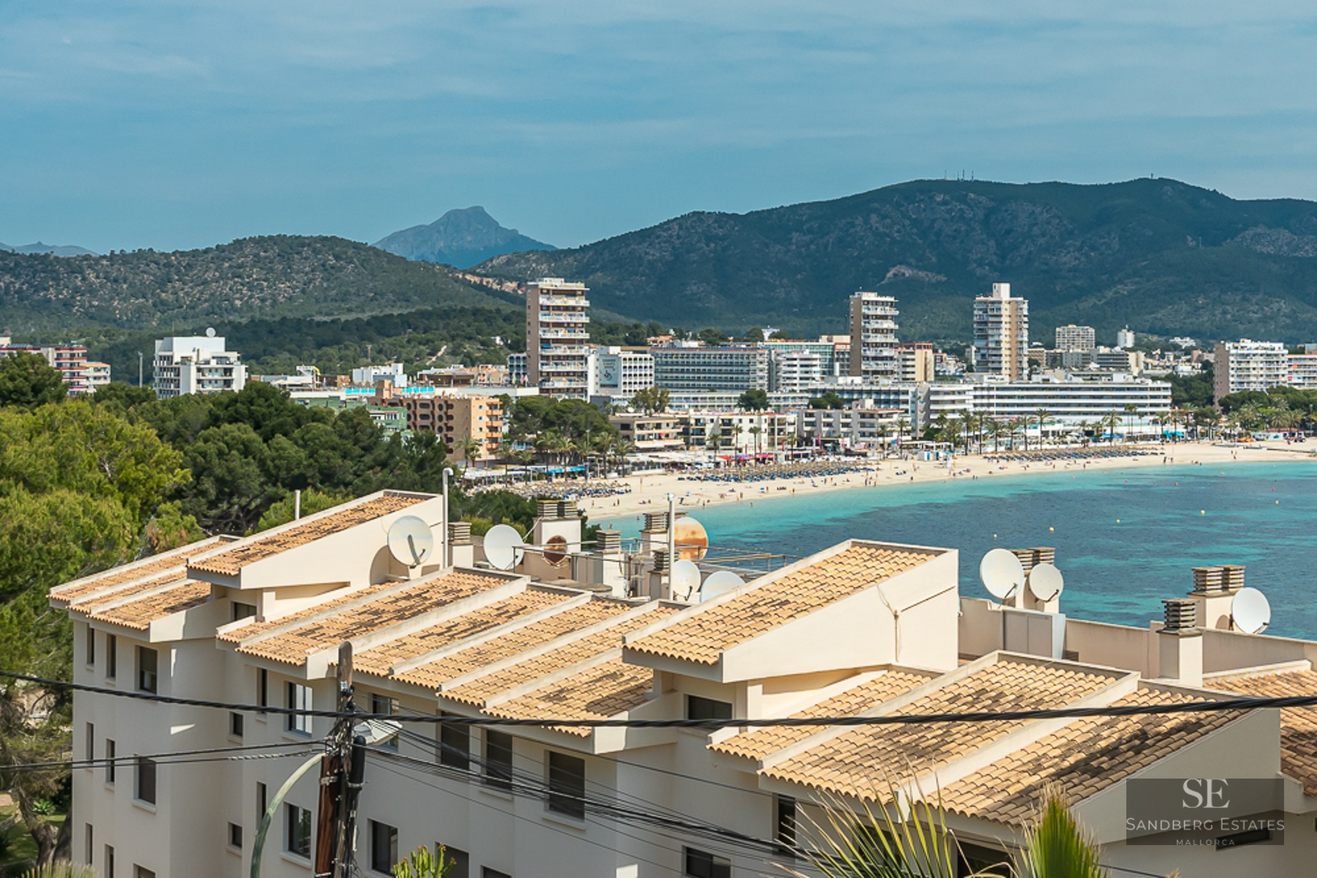 View overlooking a coastal bay with turquoise water, a sandy beach, white hotels, and green mountains in the background.