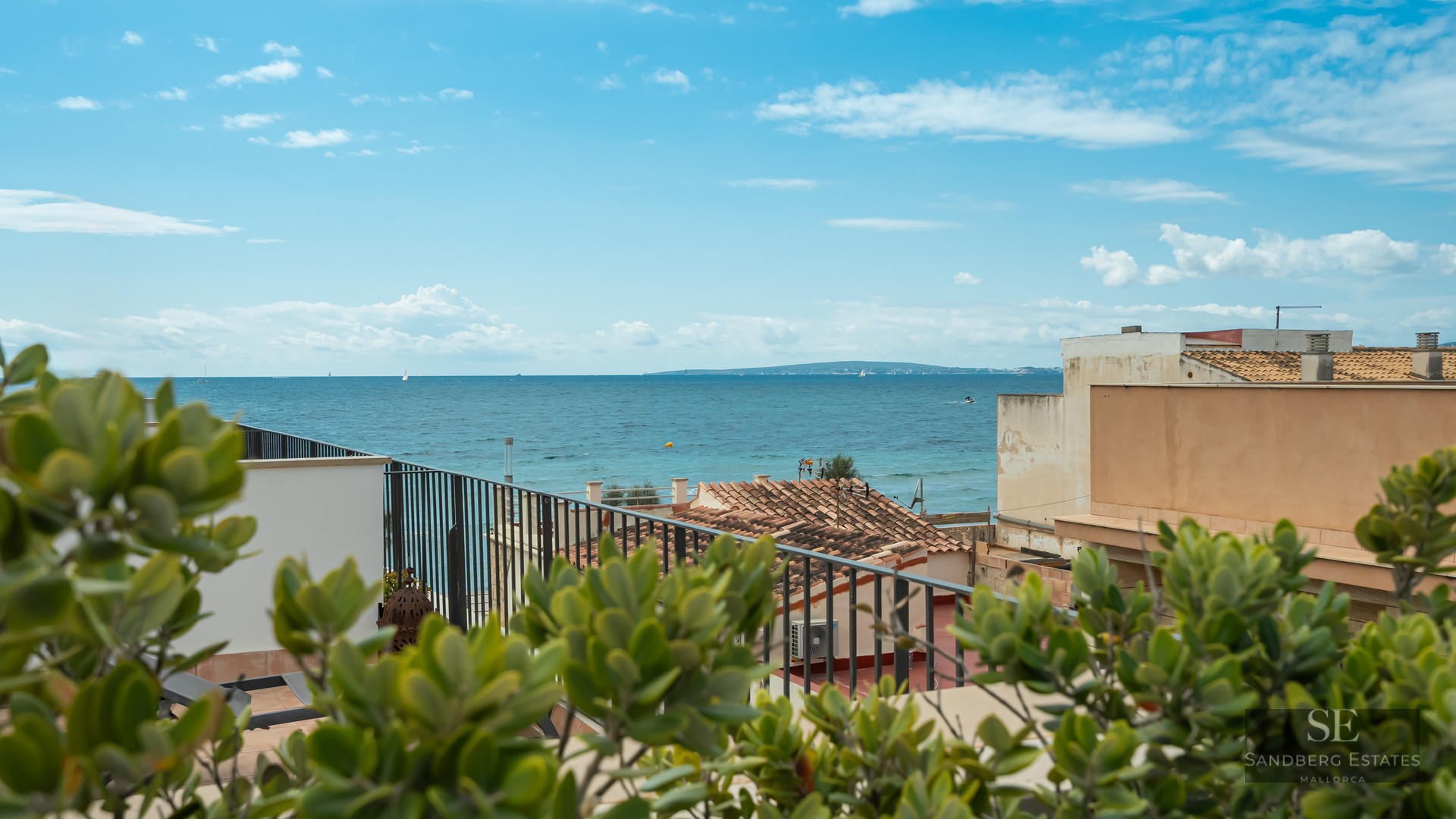 A view of the blue Mediterranean sea from a terrace with a black metal railing and terracotta roofs in the foreground.