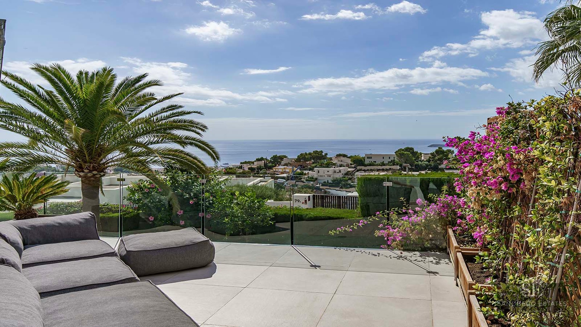 Luxury terrace with a grey outdoor sofa, glass railing, palm trees, and a stunning view of the sea and town.