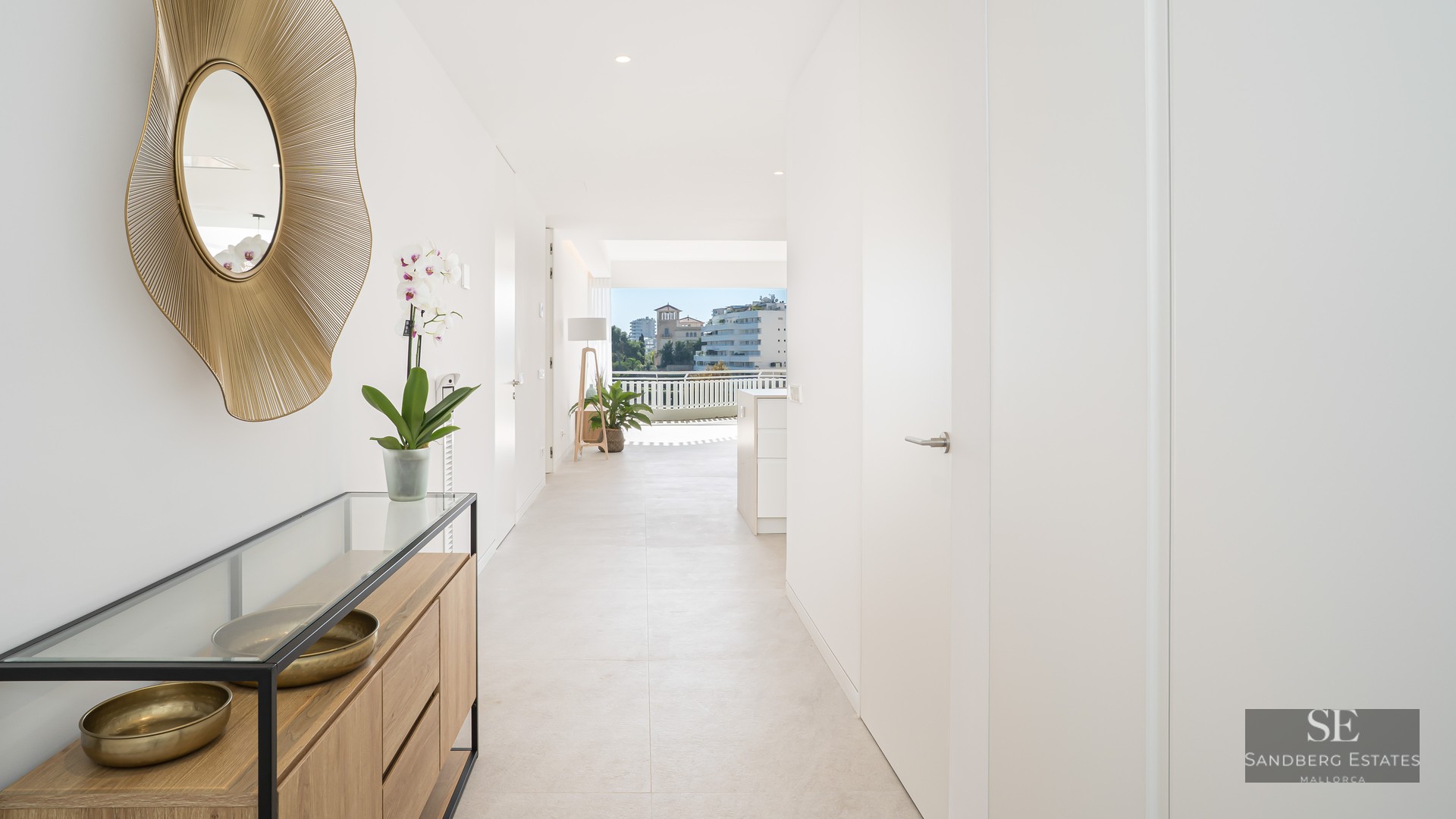 White hallway with gold sunburst mirror and wooden console, leading towards a sunny city view.