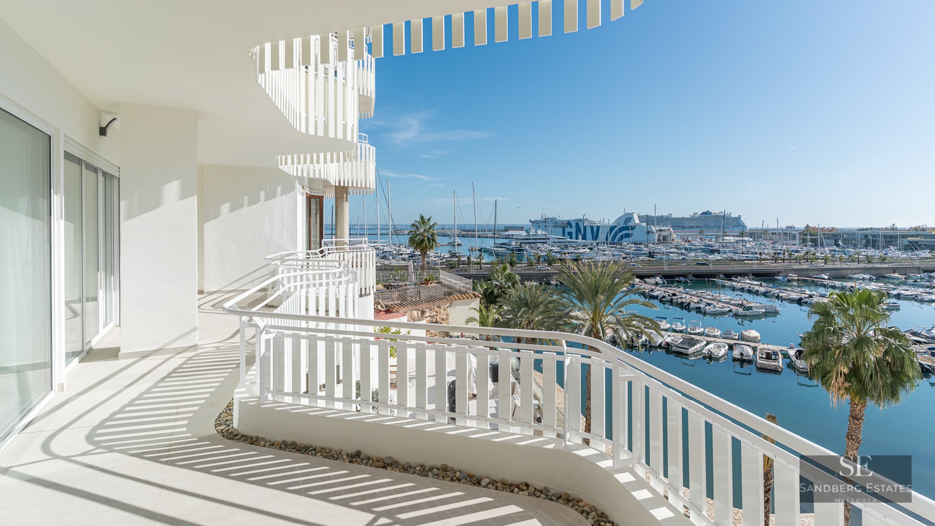 Large modern white terrace overlooking a sunny marina filled with boats and palm trees under a clear blue sky.
