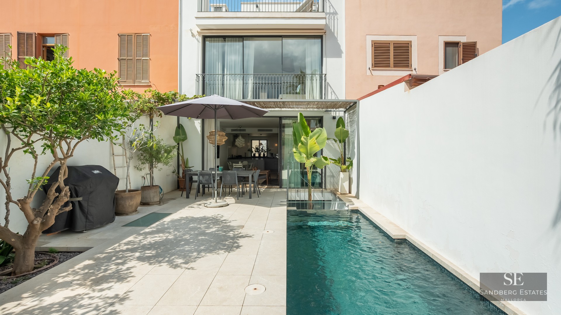 Modern outdoor courtyard featuring a lap pool, dining area under a parasol, and white walls.