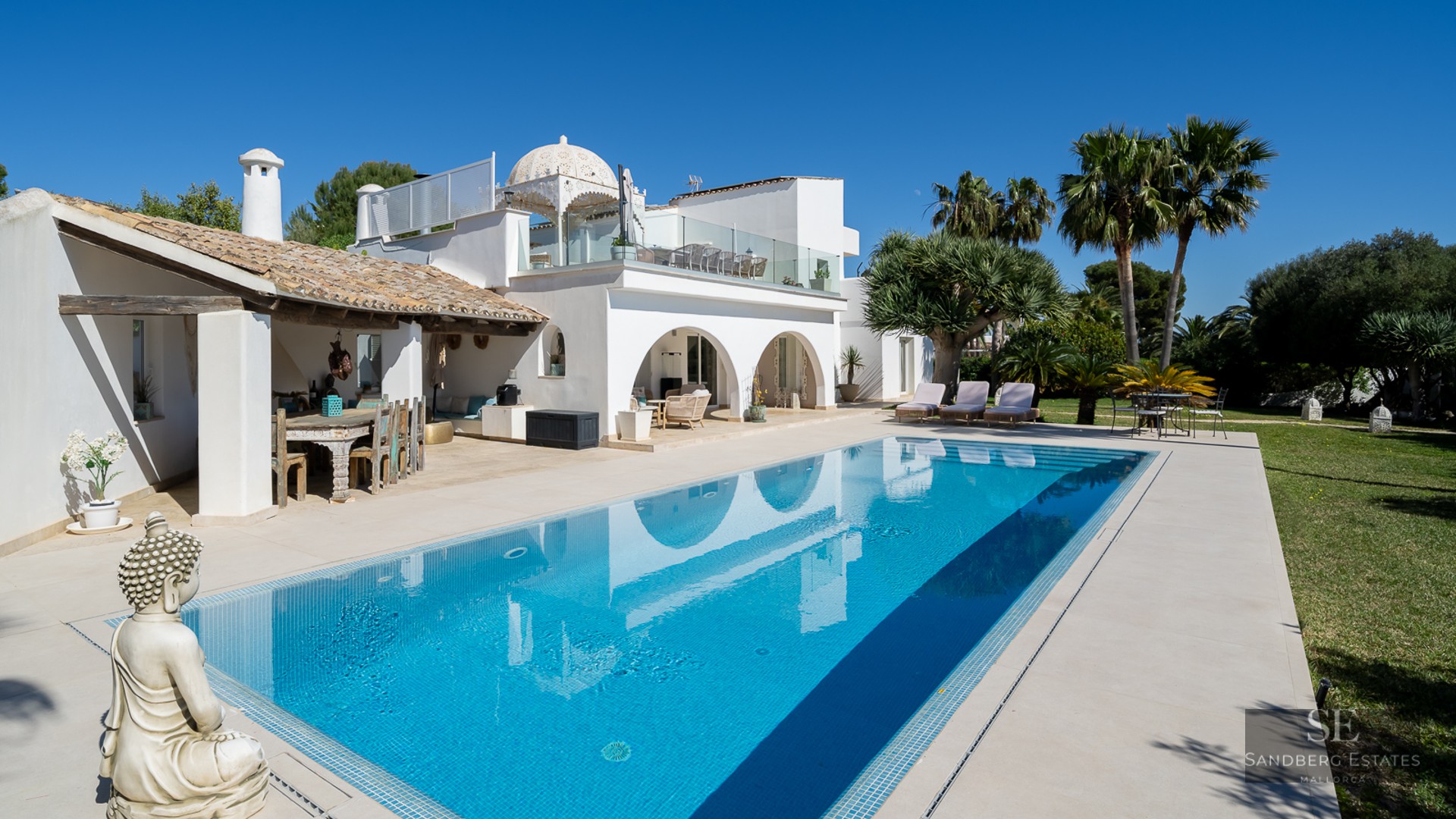 A large blue swimming pool reflecting a white Mediterranean villa with arches, palm trees, and a Buddha statue.