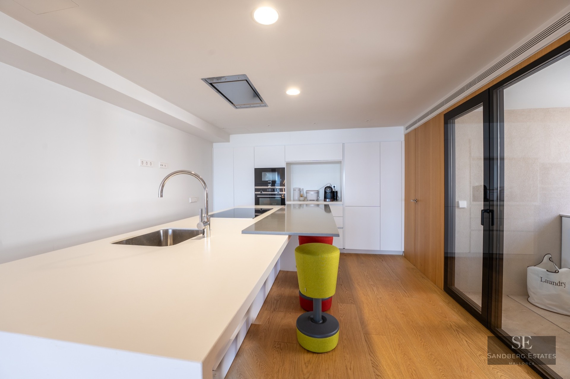Bright modern kitchen interior featuring a large white island, sink, wooden floors, and built-in appliances.