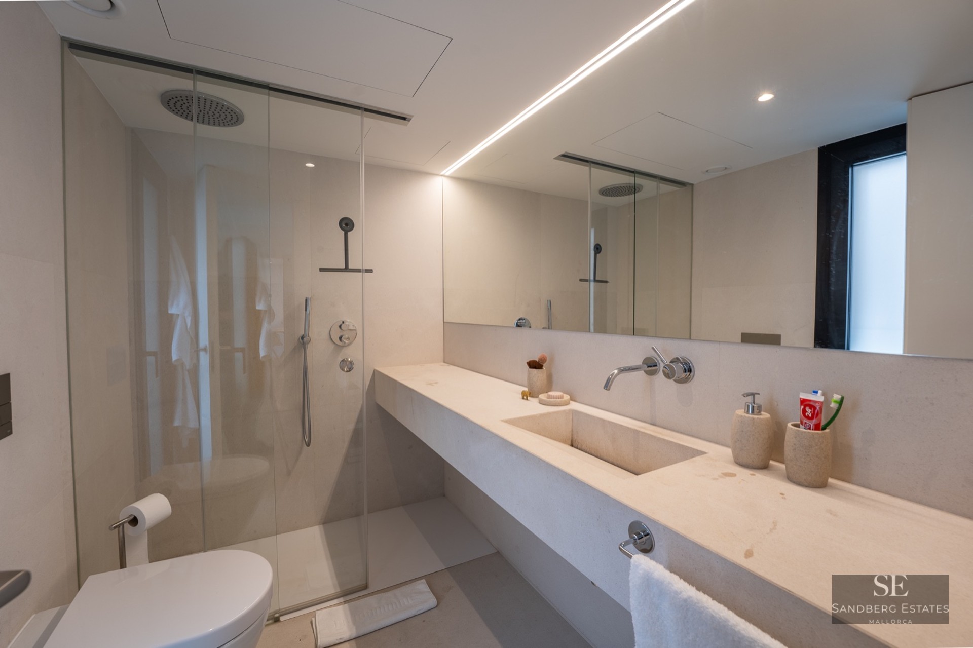 Modern bathroom with cream stone vanity, glass walk-in shower, large mirror, and linear lighting.