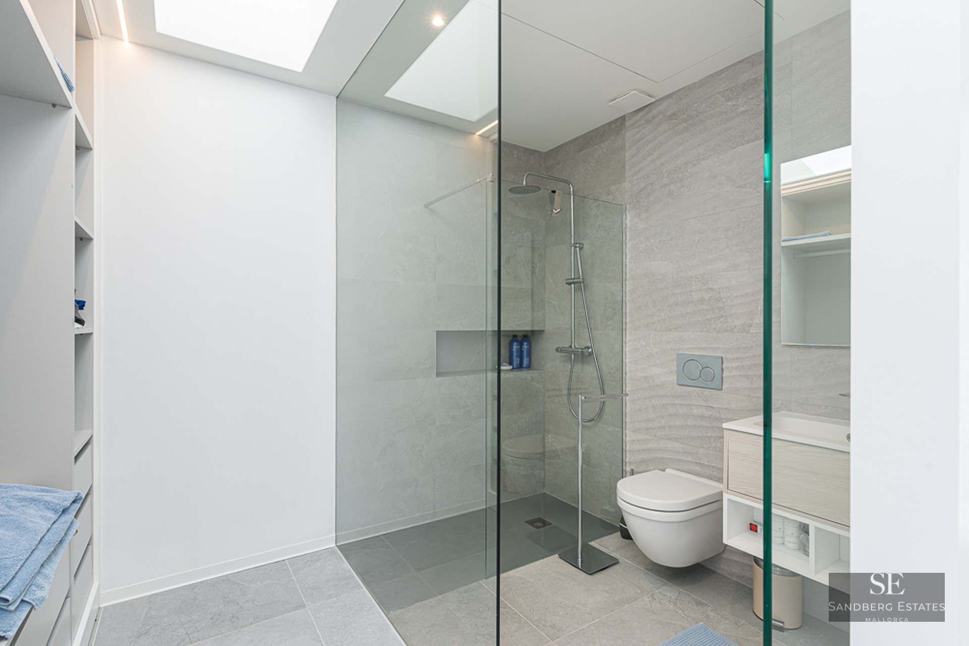 Minimalist bathroom featuring grey stone tiles, a glass walk-in shower, wall-hung toilet, and a bright skylight.