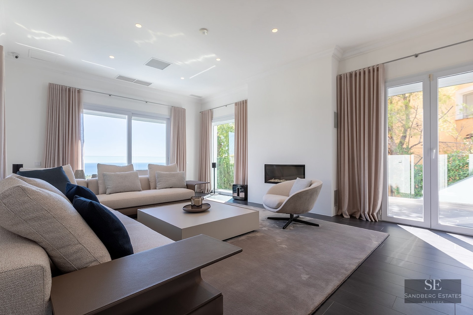 Spacious living room with cream sofas, built-in fireplace, dark flooring, and large windows overlooking the sea.