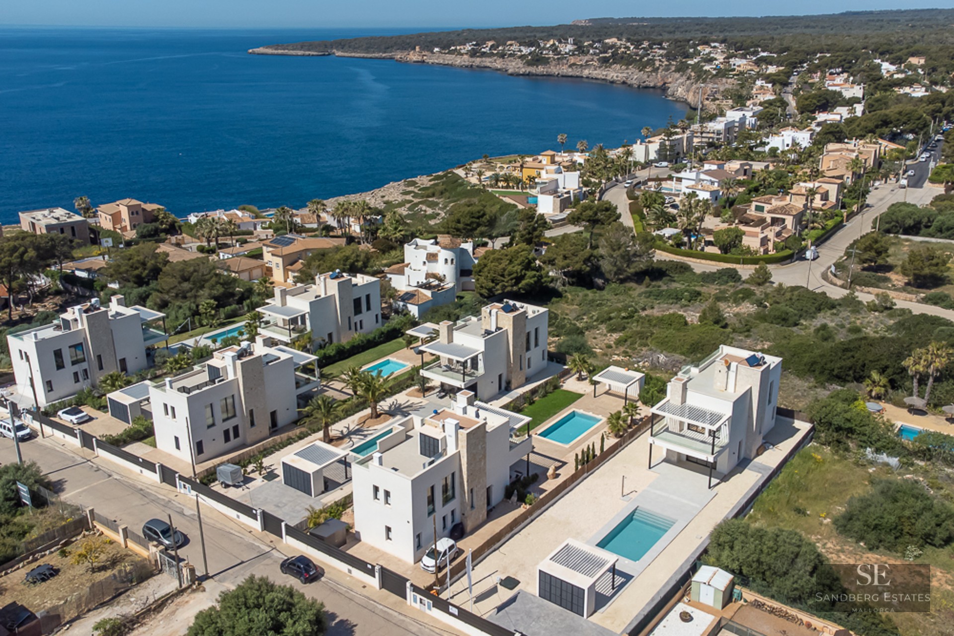 Drone view of modern white villas with private pools on a rocky coastline next to the blue sea.