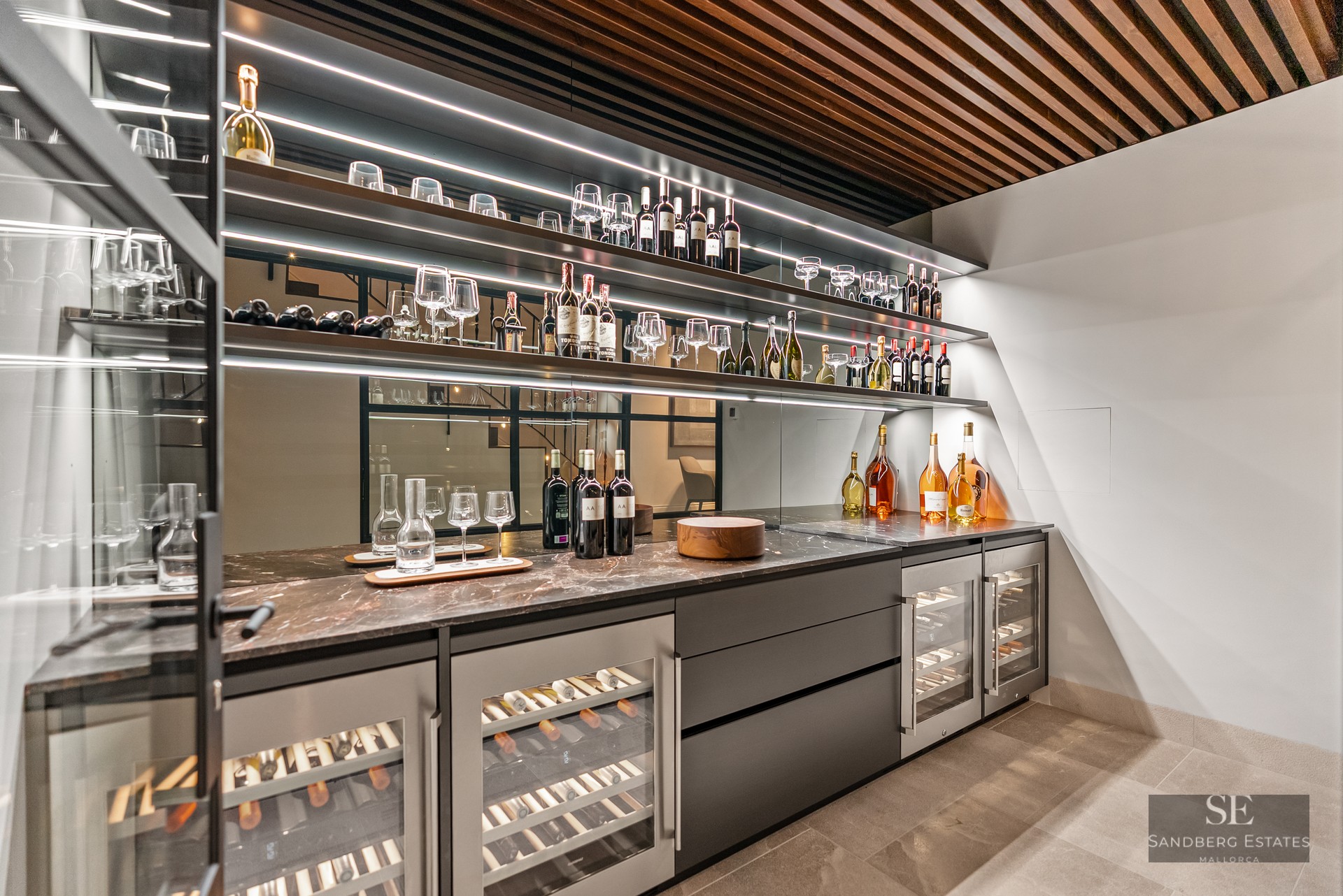 Modern wine cellar with illuminated shelves, wine coolers, marble counters, and a wooden slatted ceiling.