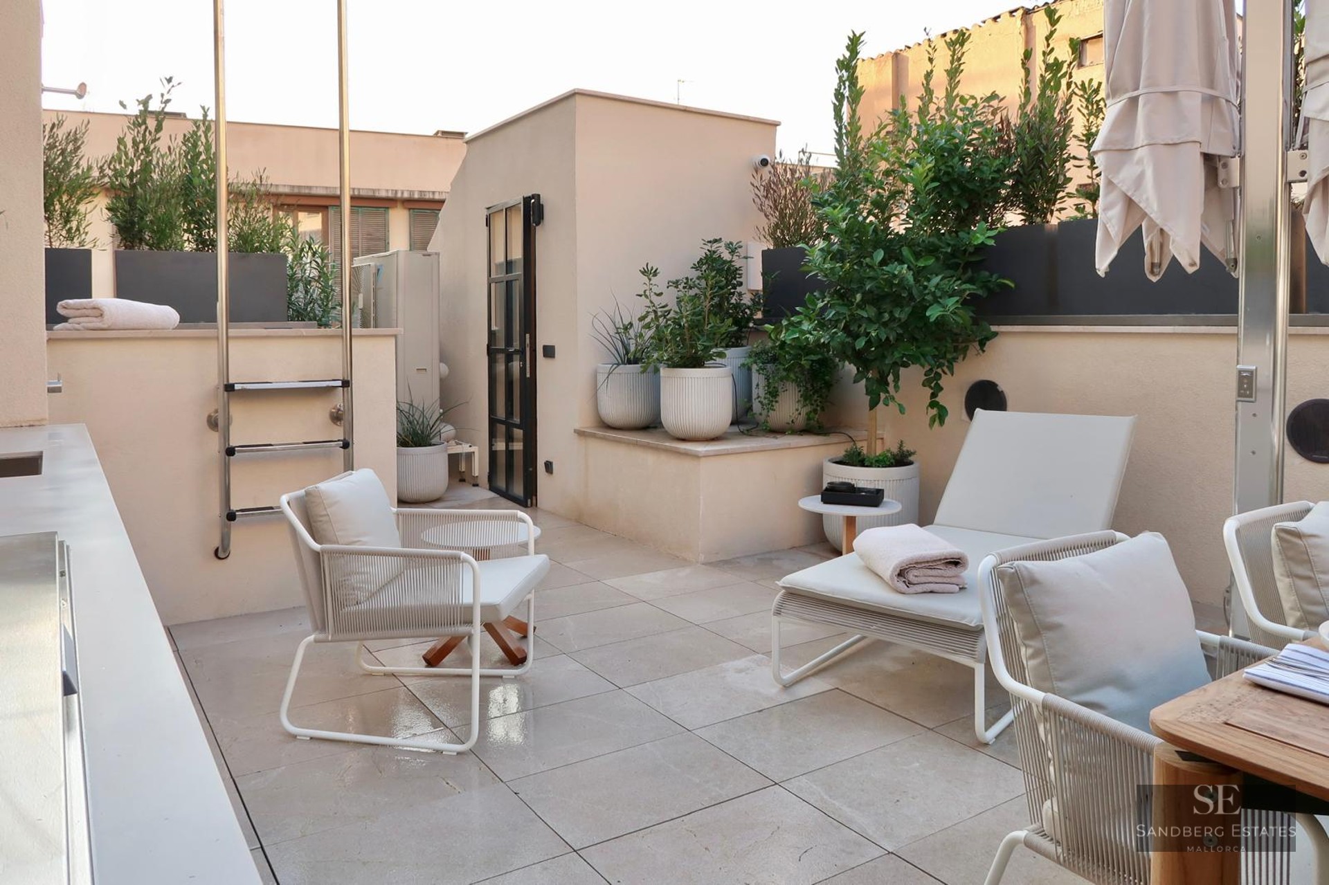 A chic rooftop terrace with white designer armchairs, a sun lounger, potted green plants, and light-colored floor tiles.