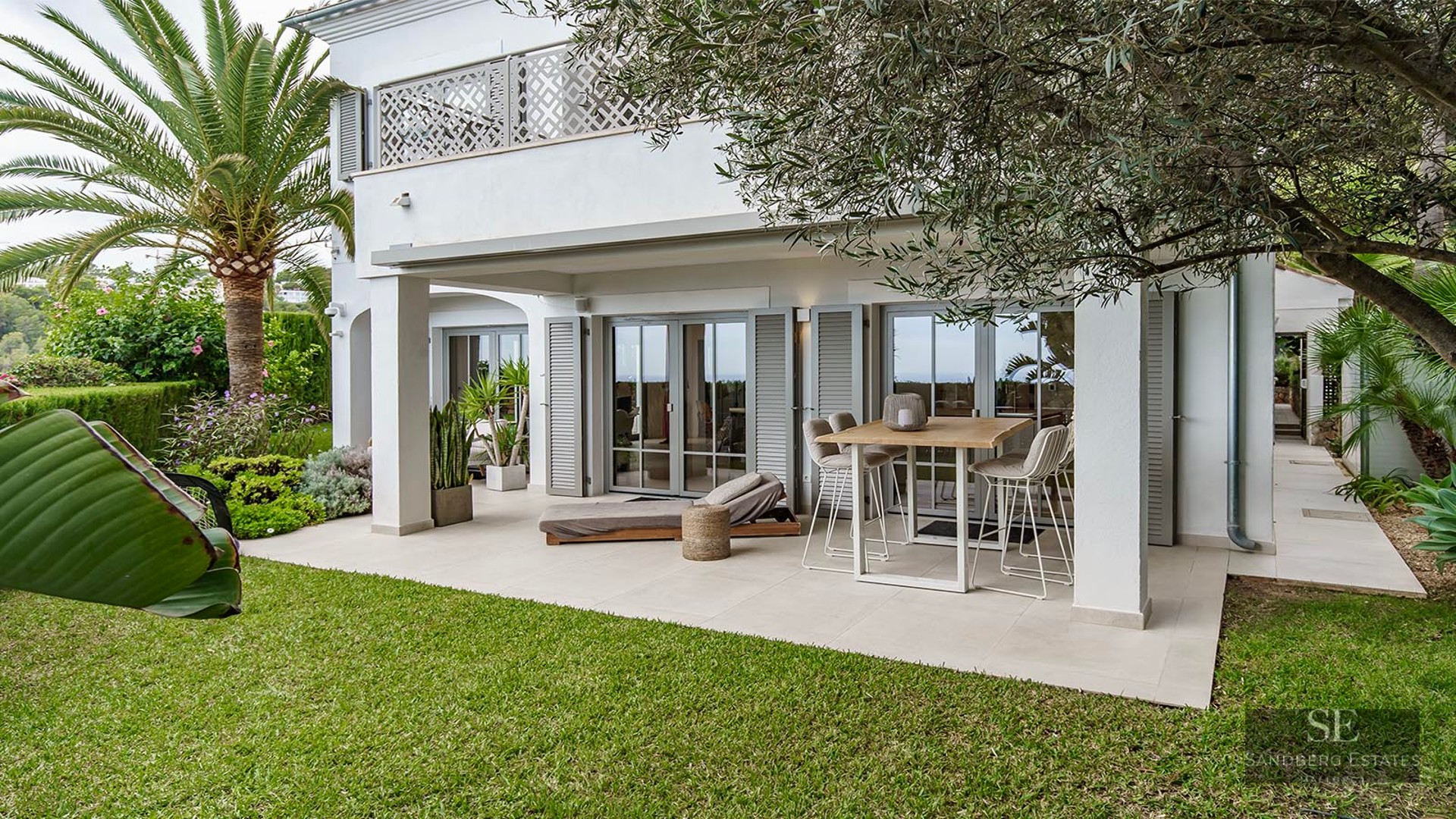 White Mediterranean villa with a covered patio, high dining table, sun lounger, green lawn, and palm trees.